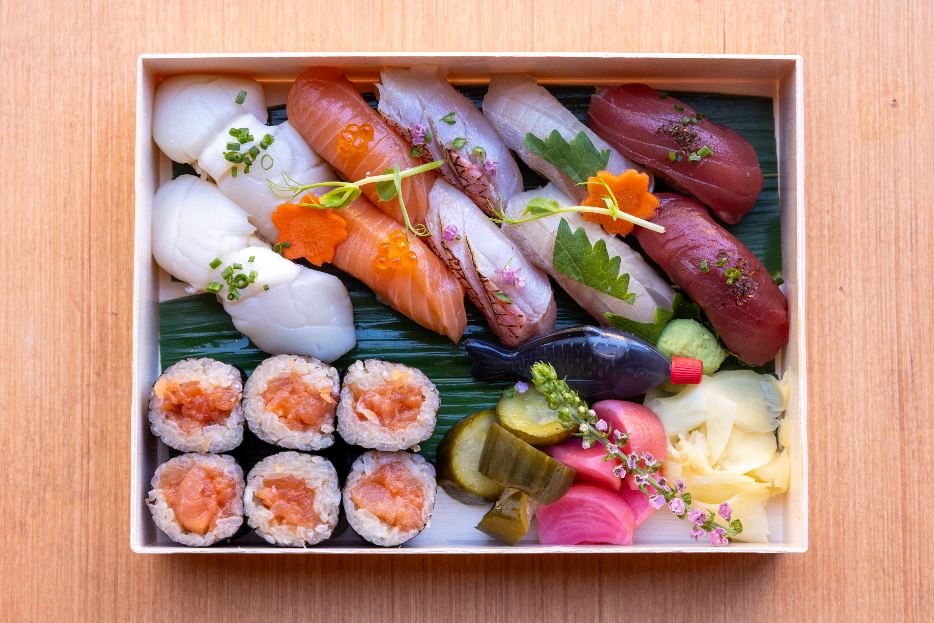 A wooden tray features assorted sushi including nigiri with scallops, salmon, and tuna, maki rolls, pickled vegetables, wasabi, ginger, and a small soy sauce container.