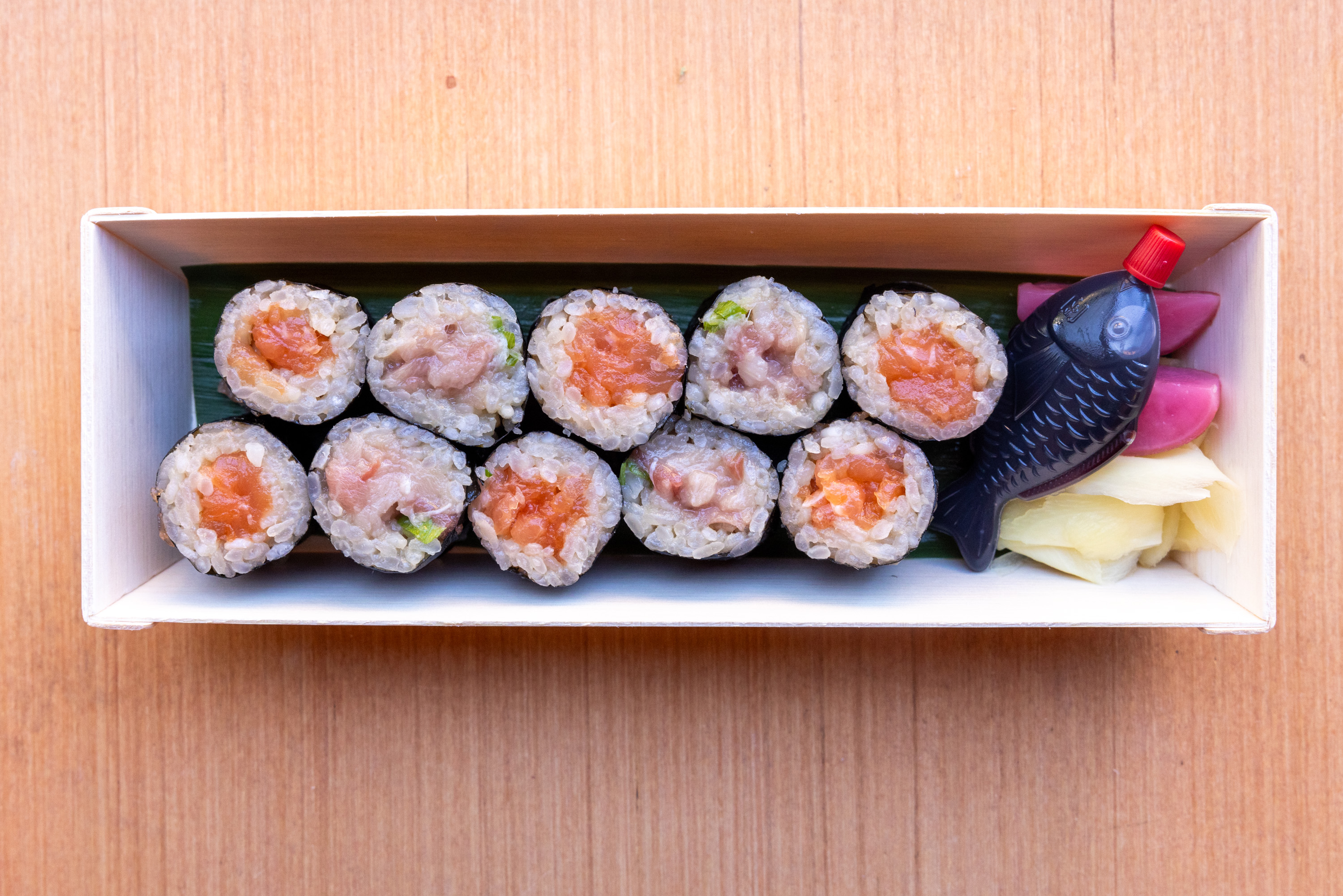 The image shows a wooden box containing eight pieces of sushi rolls, a small fish-shaped soy sauce container, and pickled ginger beside purple pickled vegetables.