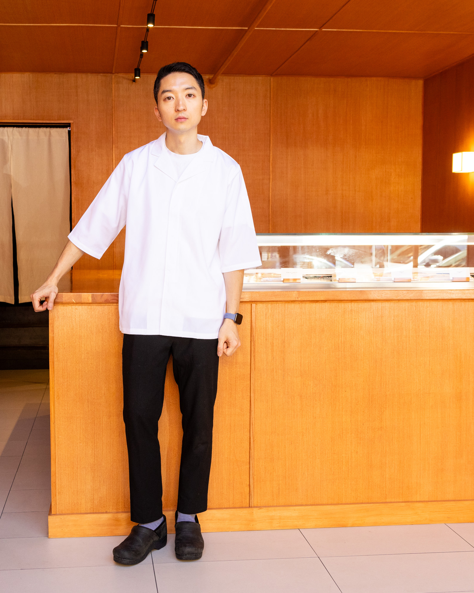 A man in a white coat stands behind a wooden counter in a minimalistic, warmly lit room, with beige curtains in the background and a display case on the counter.