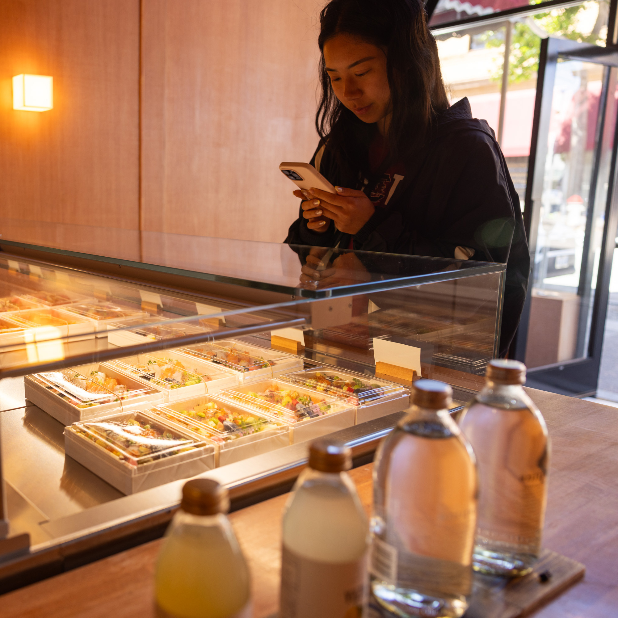 A woman stands in front of a glass display case filled with neatly arranged food containers, holding her phone. Bottles of beverages are on the counter nearby.