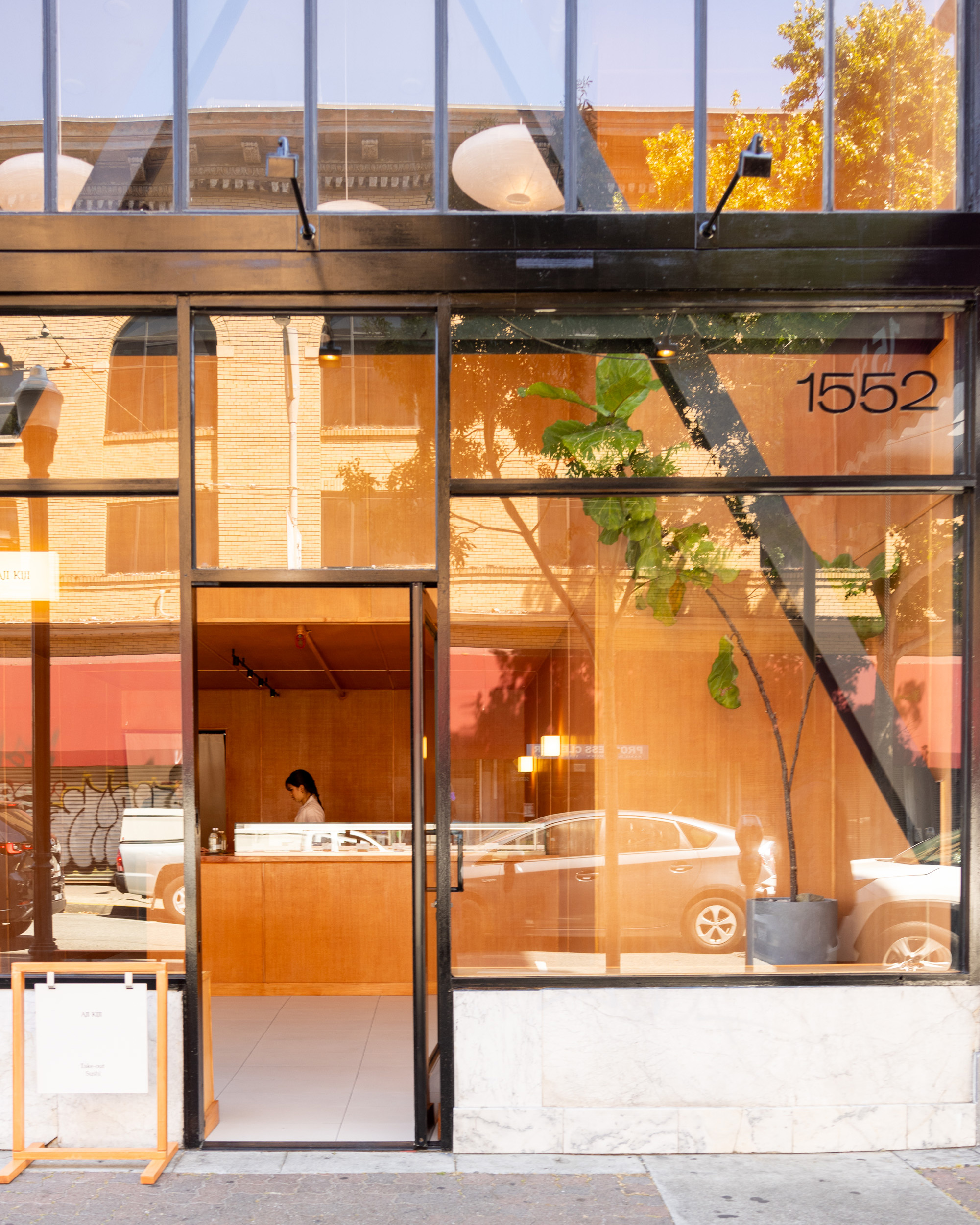 A modern storefront glass door at 1552 reveals a clean, minimalistic interior with wood paneling, a potted plant, and a person standing behind a counter inside.