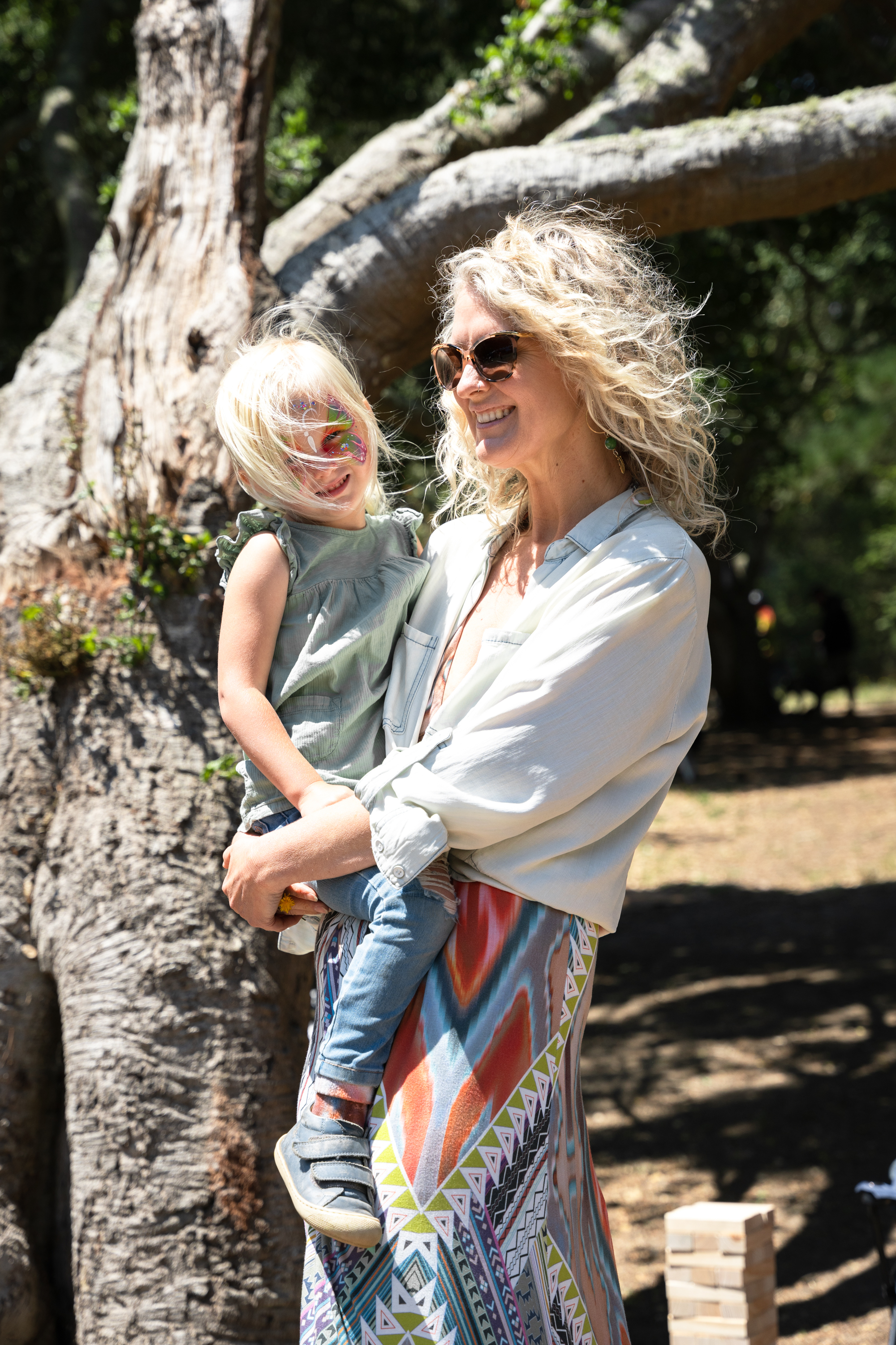 A woman with curly hair and sunglasses holds a young child with face paint in front of a large tree, both are smiling and dressed in casual, colorful clothing.