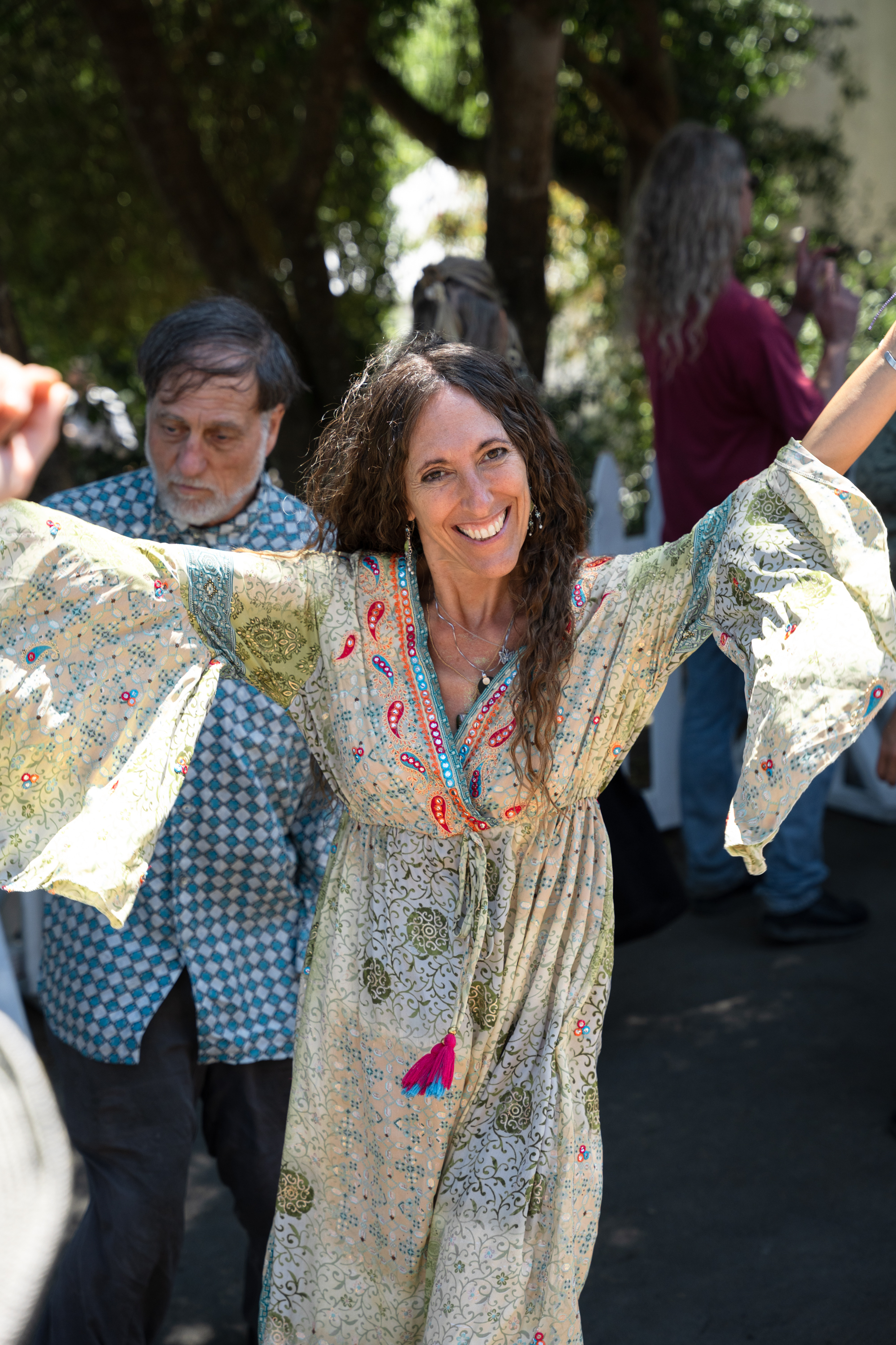 A woman with long curly hair smiles brightly and spreads her arms cheerfully in an outdoor setting. She wears a long, patterned dress with bell sleeves. Others are visible in the background.