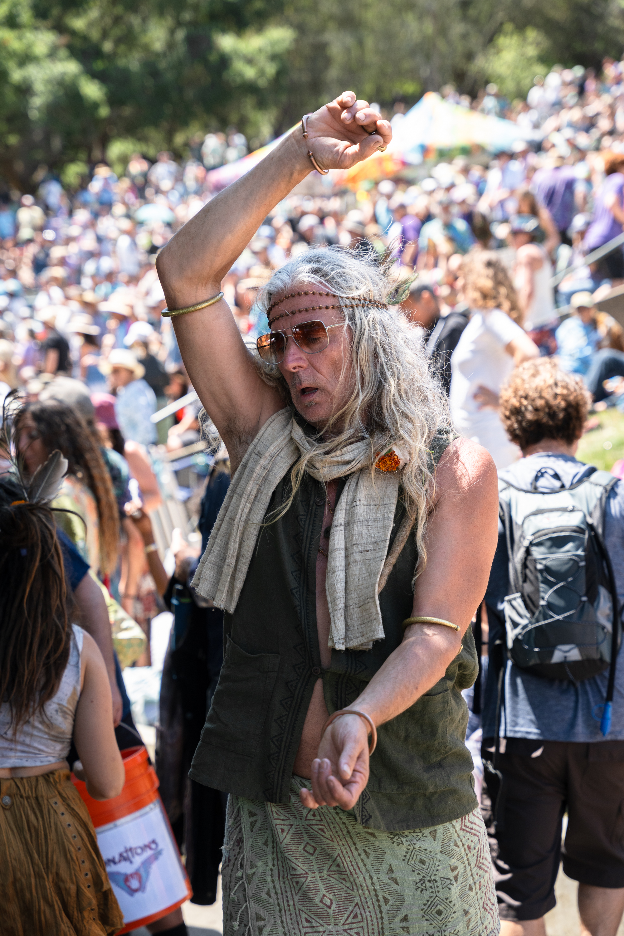 A person with long gray hair, wearing sunglasses and a headband, dancing among a large, seated crowd outdoors. The crowd is dense and the background is leafy.