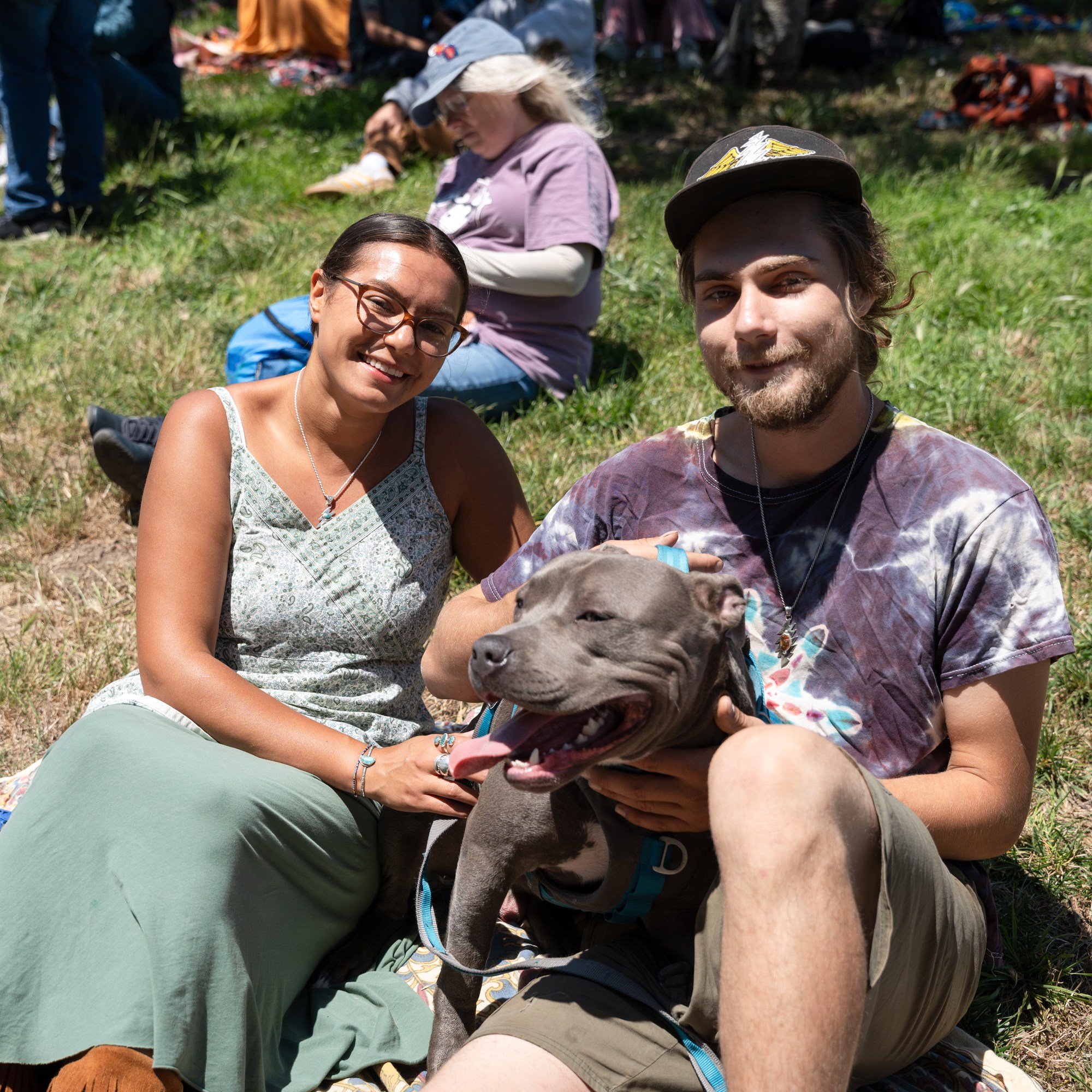 A smiling woman and man sit on a blanket in a grassy area with a happy, panting dog on the man’s lap. People and blankets are visible in the background.