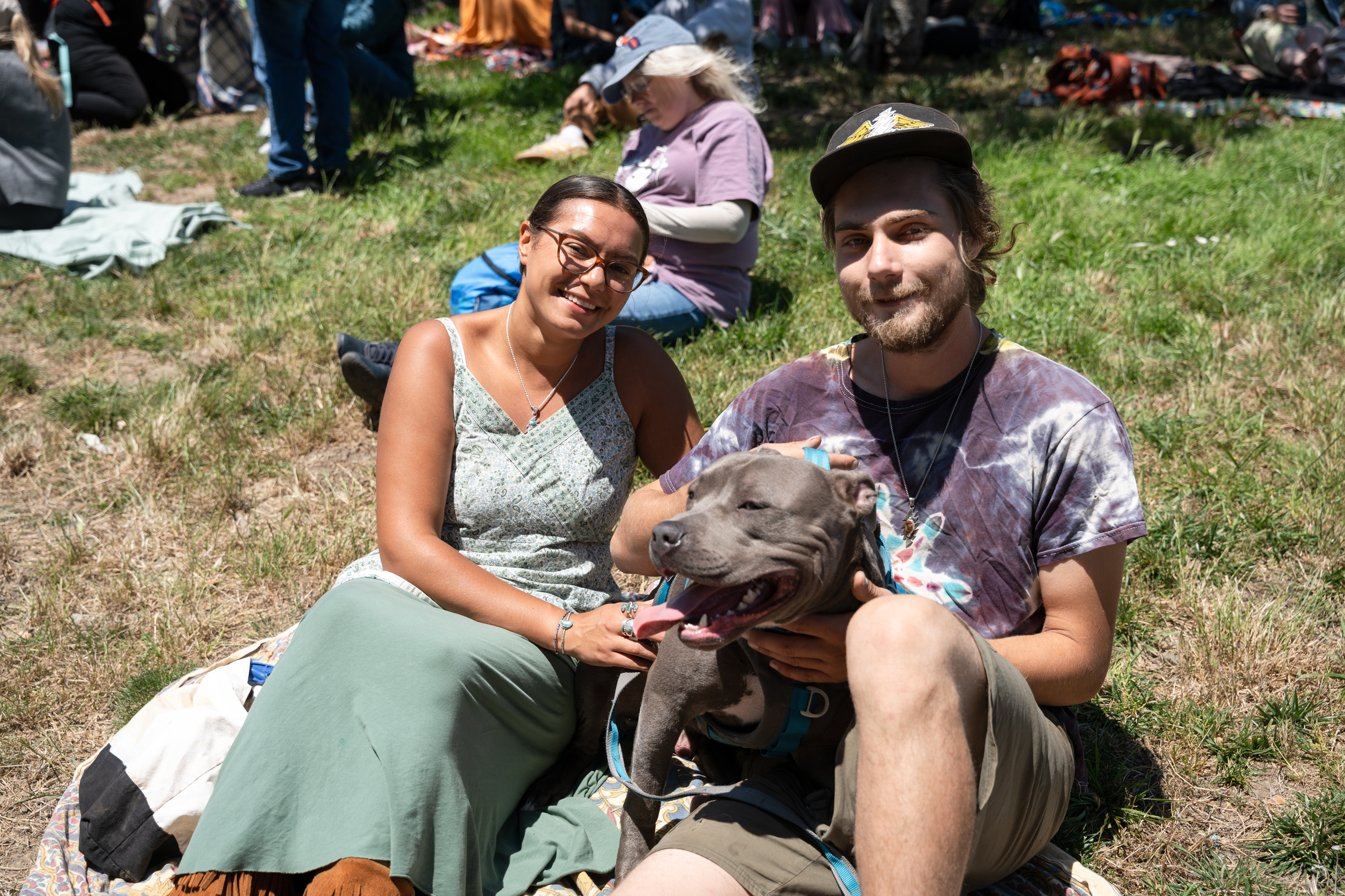 A smiling woman and man sit on a blanket in a grassy area with a happy, panting dog on the man’s lap. People and blankets are visible in the background.