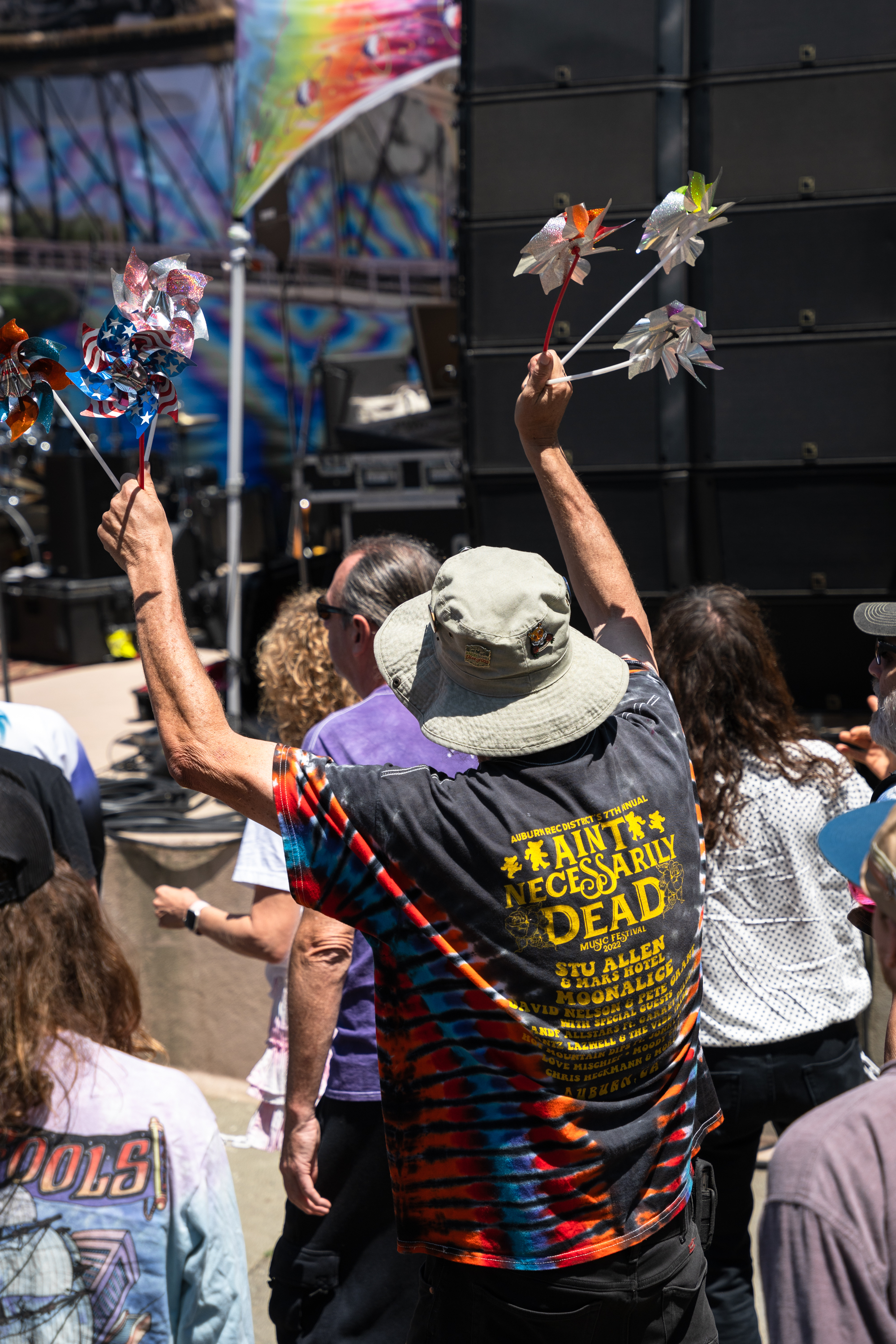 A person wearing a tie-dye &quot;Ain't Necessarily Dead&quot; shirt and bucket hat raises pinwheels in a crowded outdoor event adorned with colorful banners.