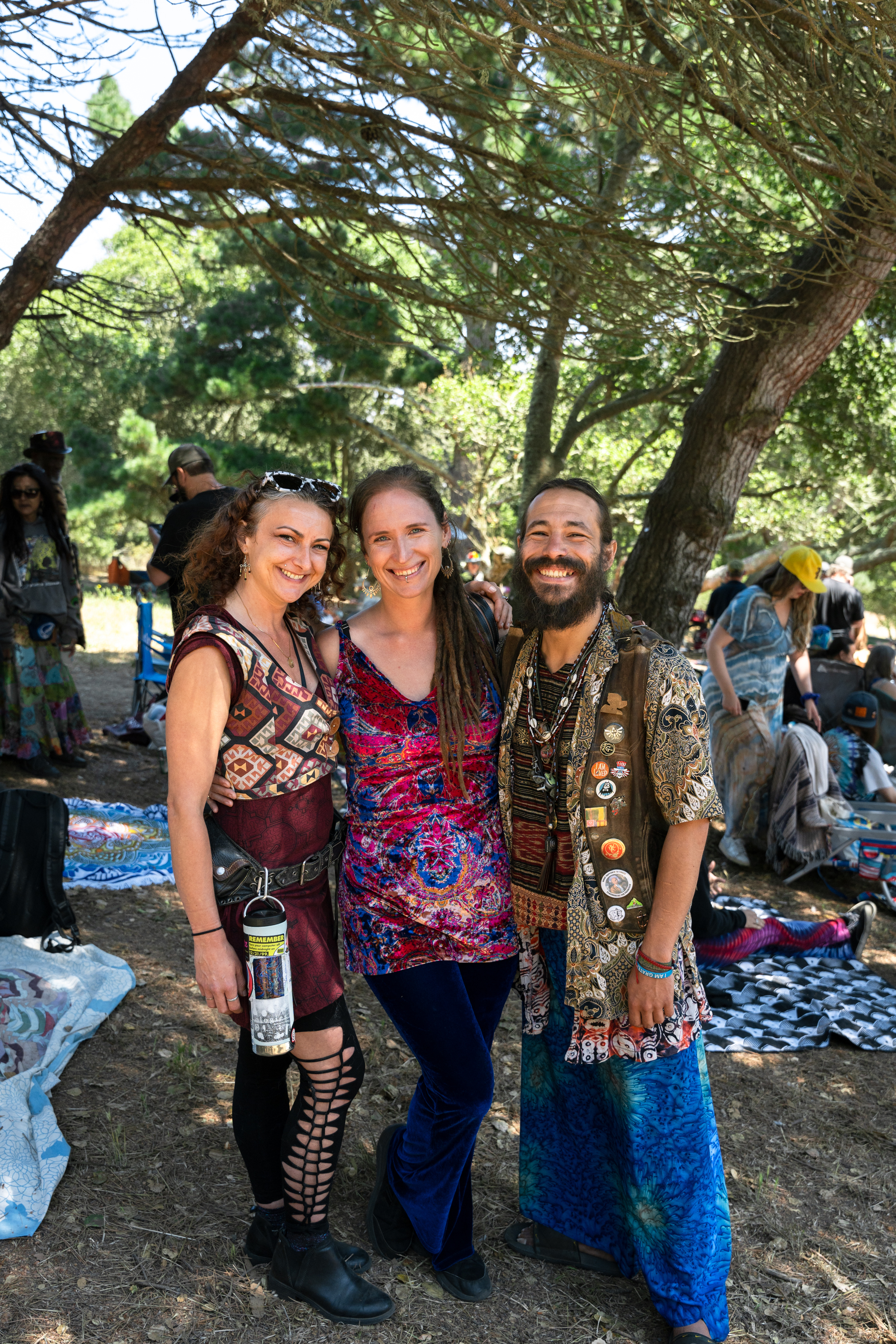 Three people are smiling and posing together under tree branches. They are dressed in colorful, eclectic clothing, and other people are gathered in the background.