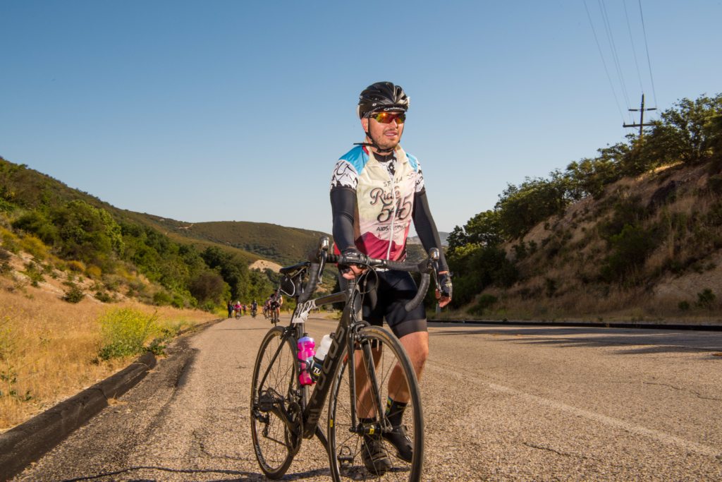 A cyclist in protective gear stands beside a bike on a rural road with hills and trees in the background; several other cyclists can be seen in the distance.