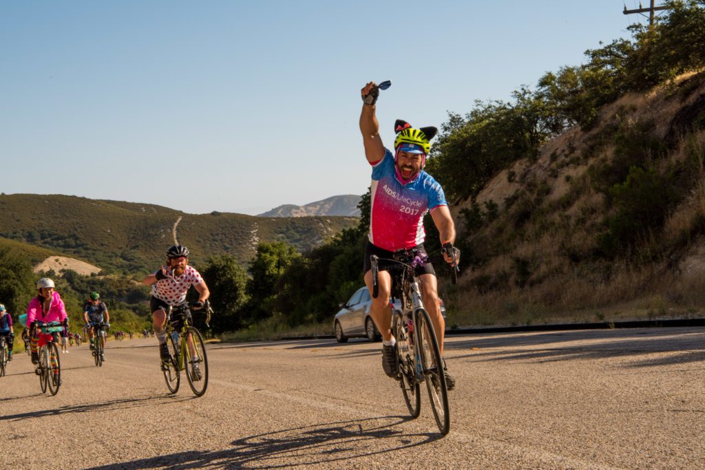 Cyclists ride along a highway in hilly terrain. One rider in front raises a hand triumphantly while others follow in the background. It is a sunny, clear day.