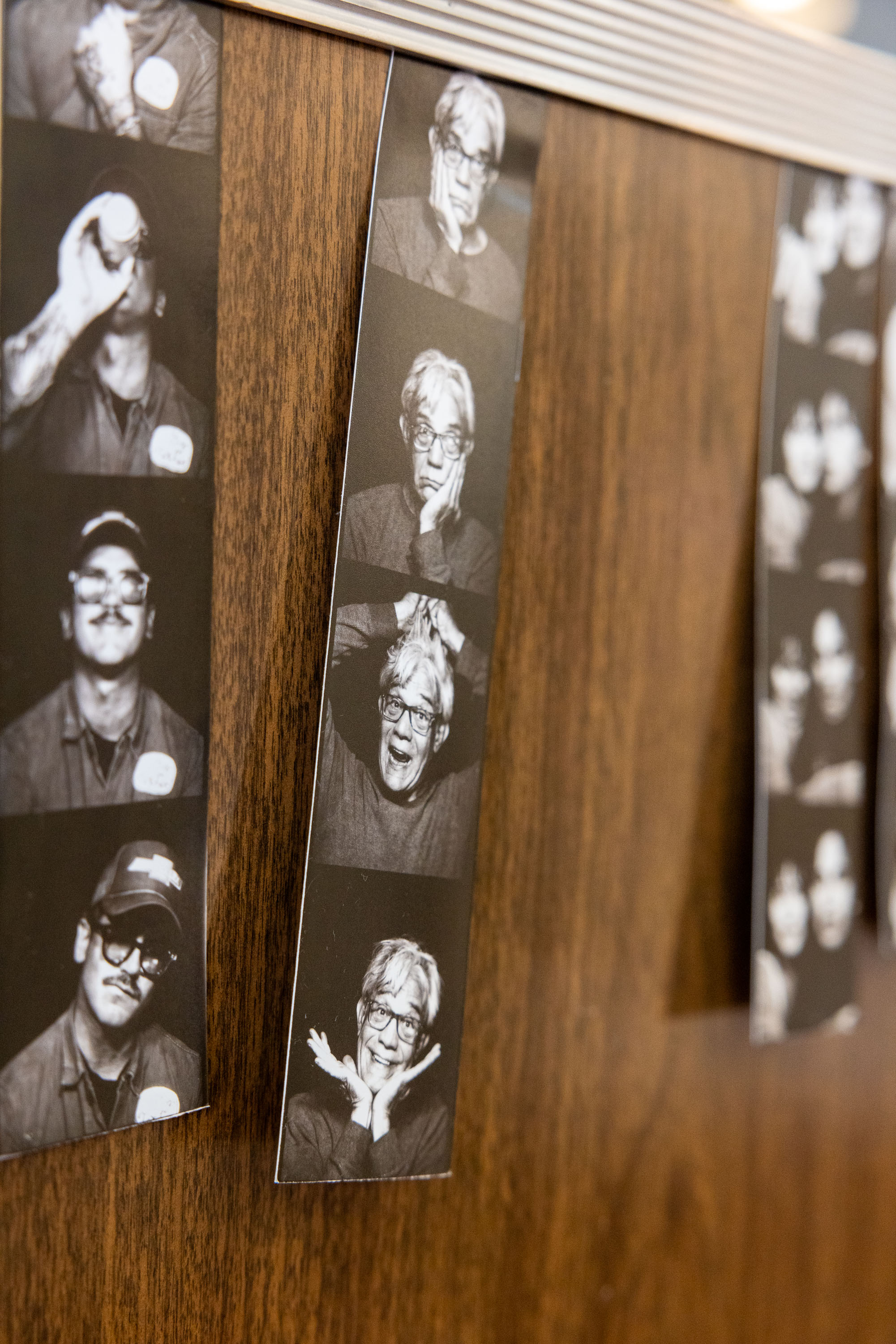 Black-and-white photo booth strips are displayed on a wooden surface. Each strip features a person making various facial expressions and poses.