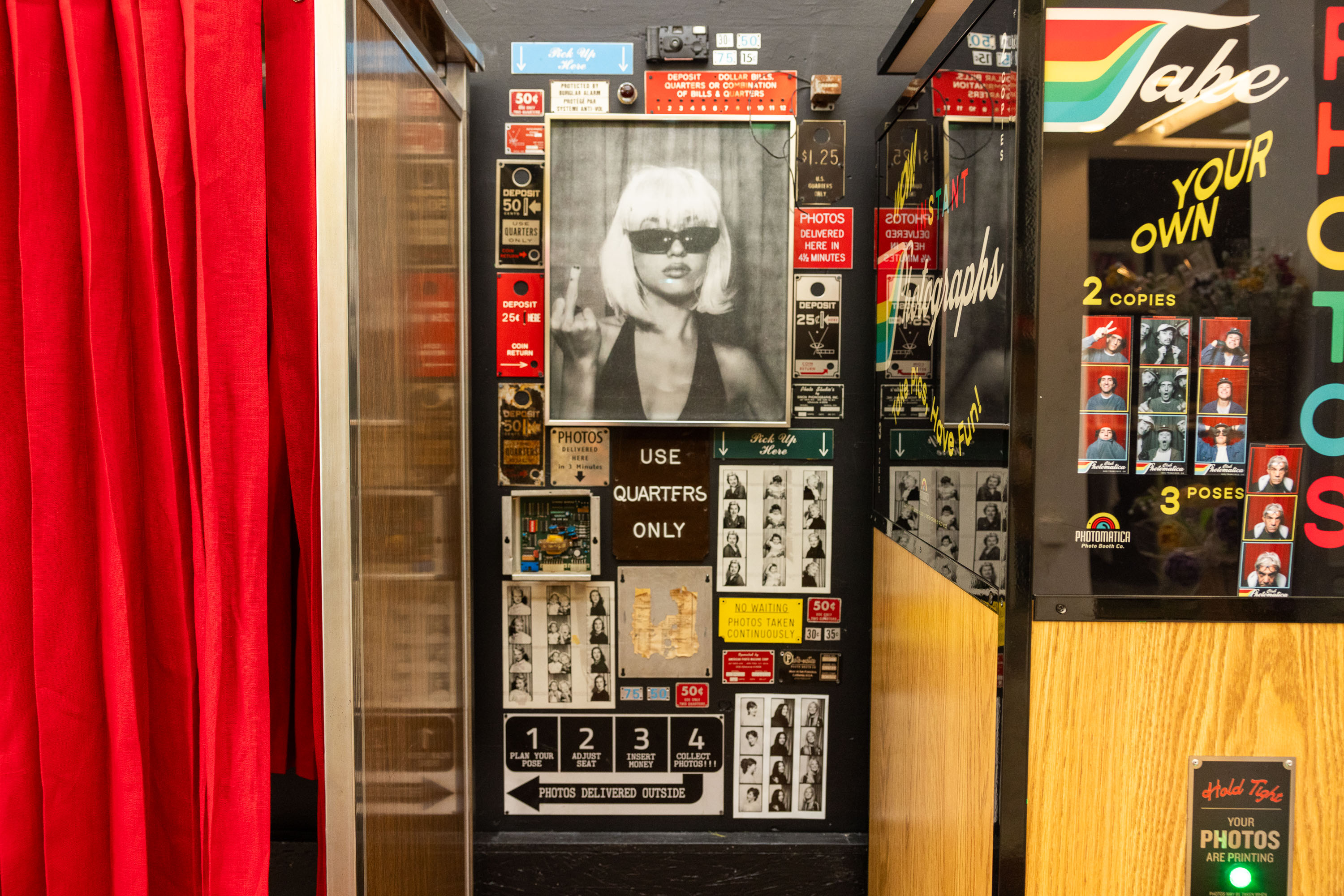 The image shows an old-fashioned photo booth decorated with various signs and photo samples, including a large picture of a blonde woman in sunglasses. A red curtain is partially drawn.