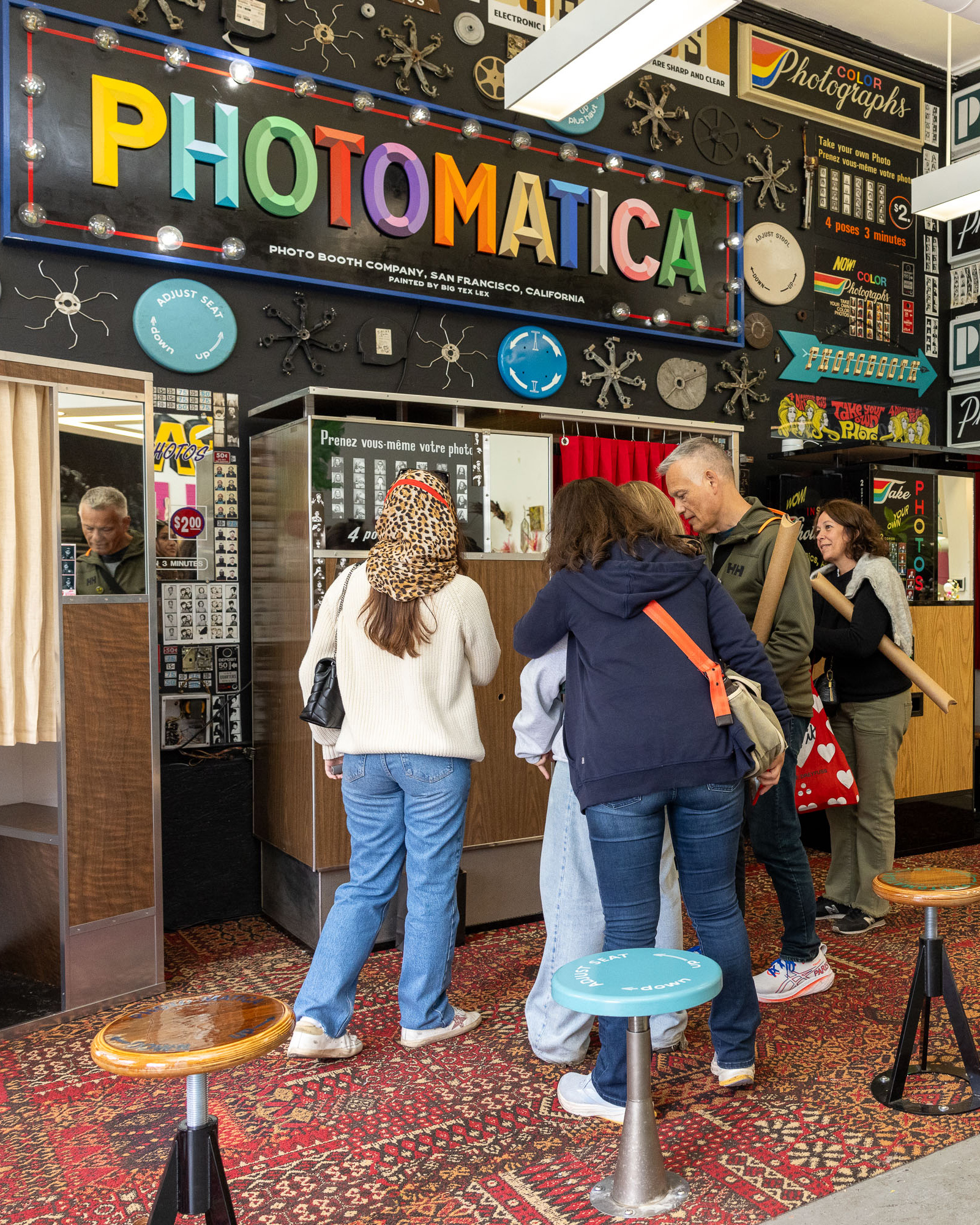 A group of people stand around a vintage photo booth labeled &quot;PHOTOMATICA,&quot; with a colorful retro sign. The interior features a patterned carpet and retro decor.