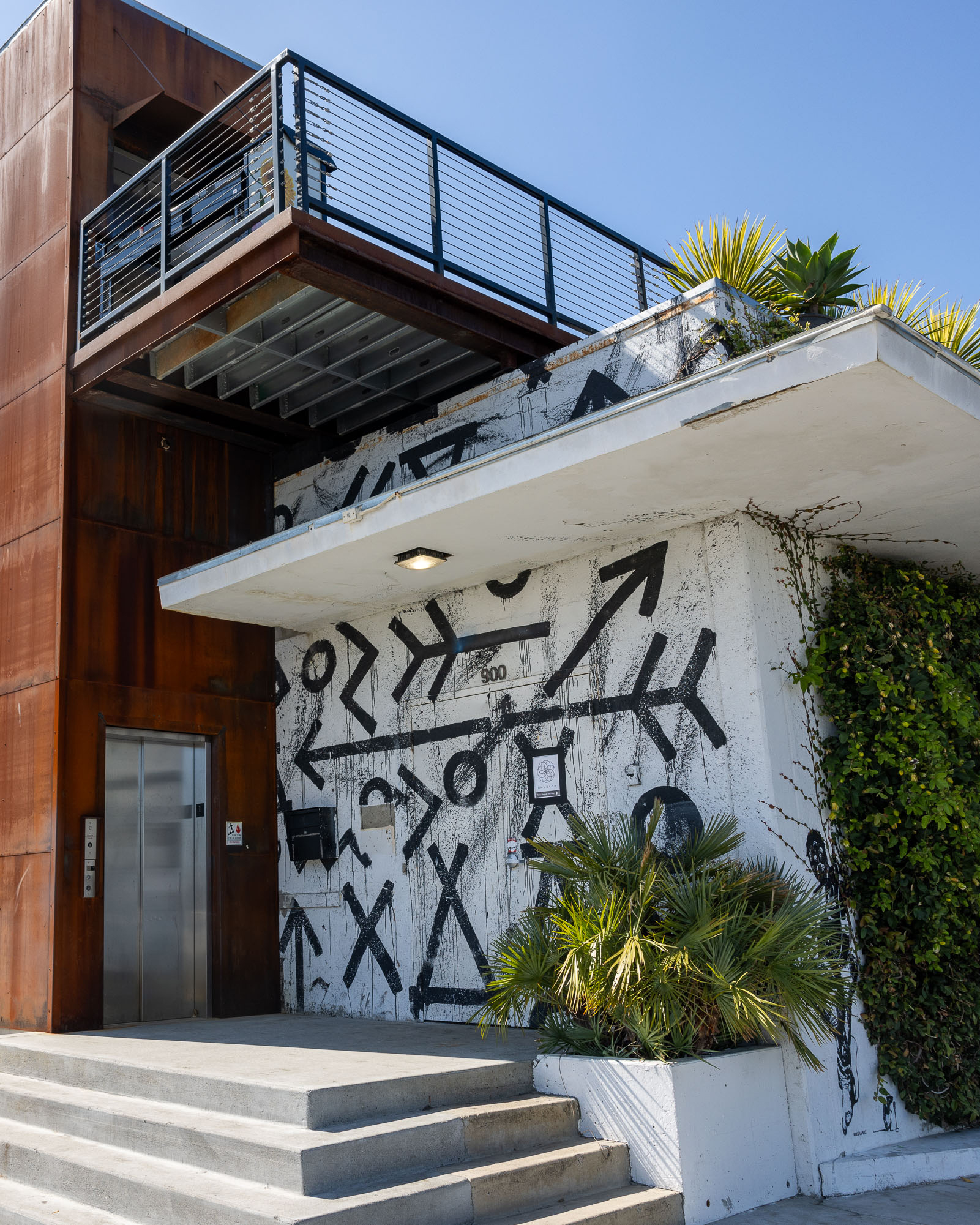 The image shows a modern building with a metal exterior, a balcony, and stairs leading to a door. The wall is adorned with bold, black abstract graffiti.