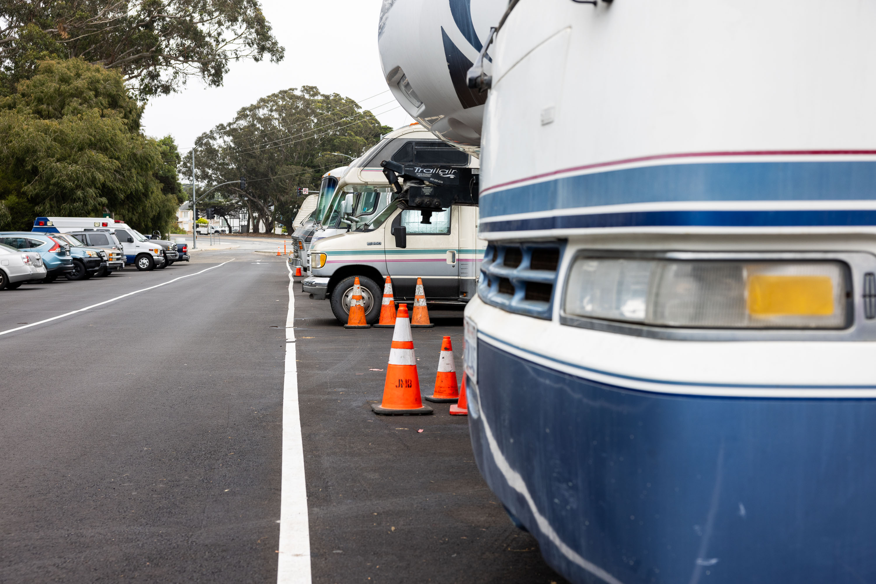 The image shows a row of parked RVs along a street. Orange traffic cones are placed near the RVs, and several cars are parked on the opposite side. Trees are visible in the background.