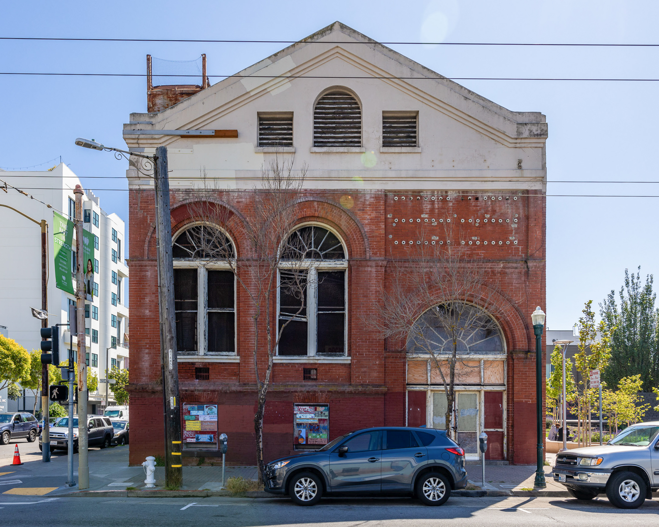 An old, two-story brick building with arched windows, boarded-up entrances, and a weathered exterior sits next to a street with parked cars and some trees.