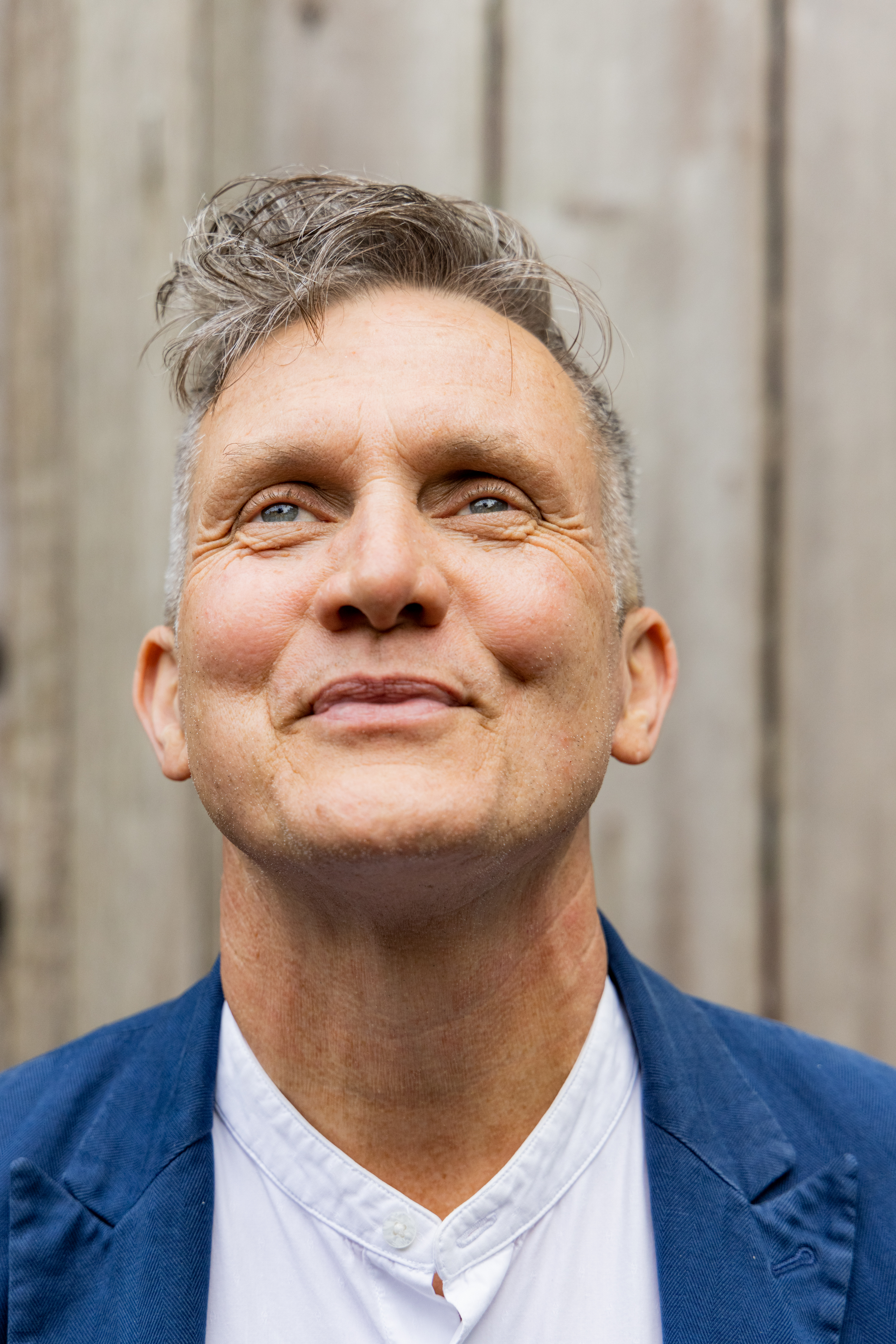 A smiling person with short grayish hair in a blue blazer and white shirt looks upwards. The background is a blurred wooden wall.