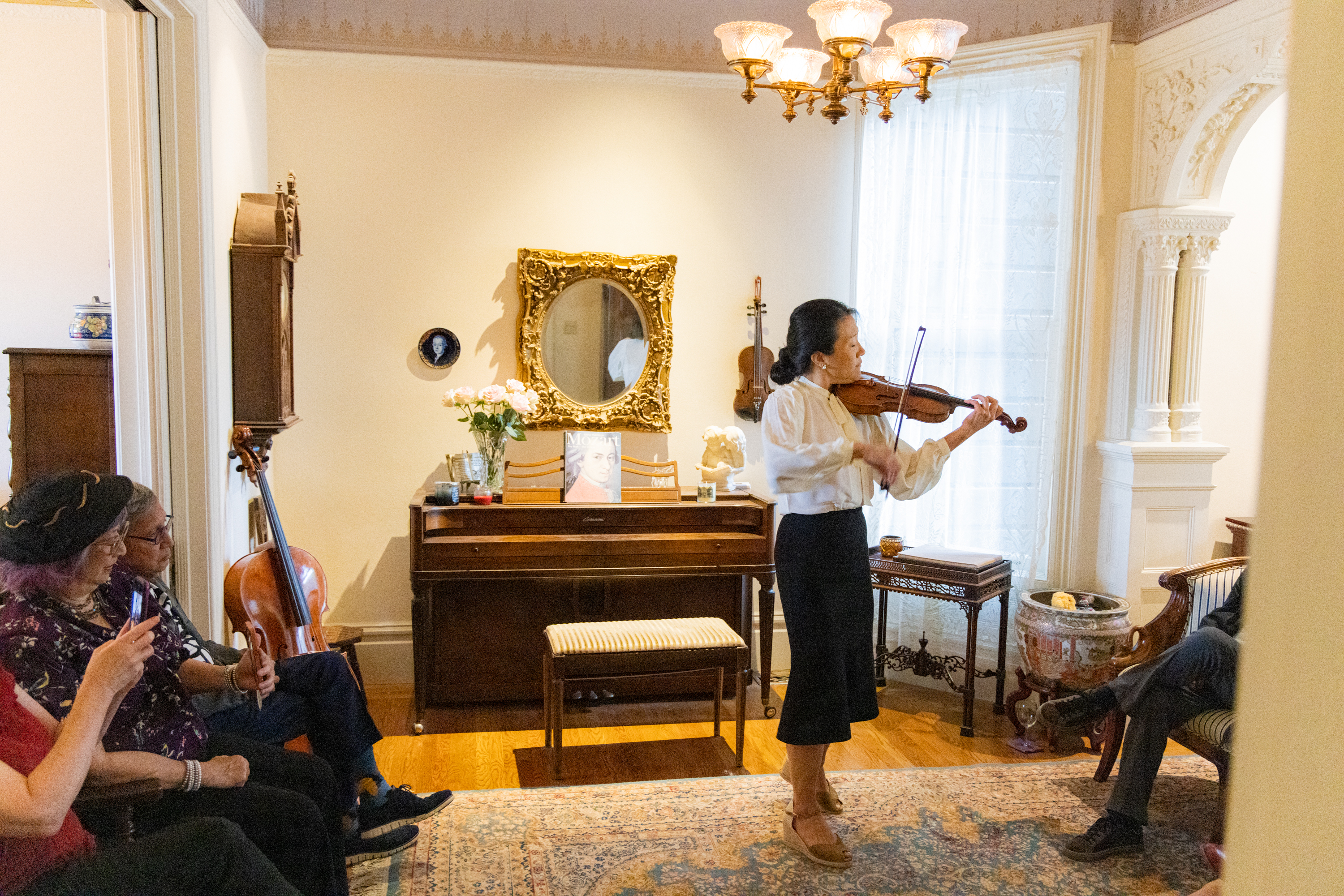 A woman in a black skirt and white shirt plays violin in an elegant living room to a small crowd. 