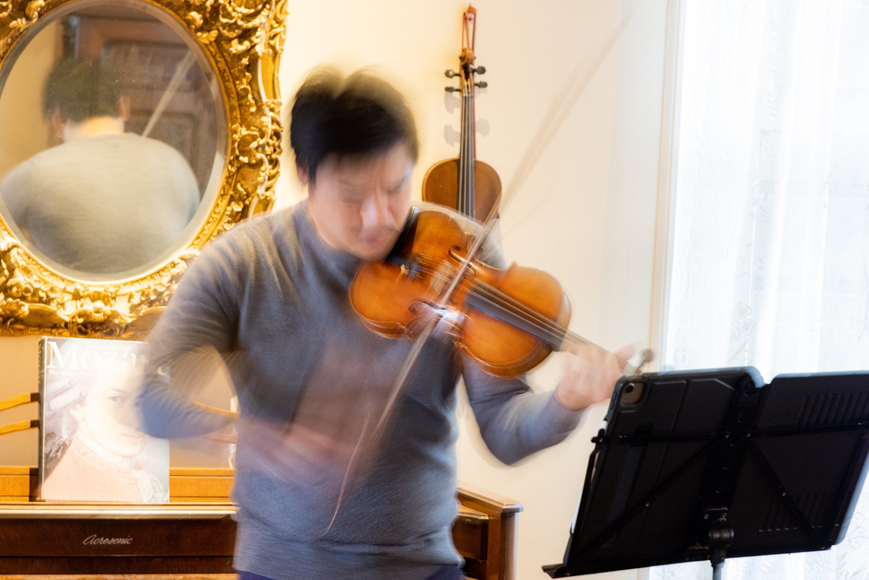 A man with a grey sweater and black hair plays a violin at great speed in an elegant room. 