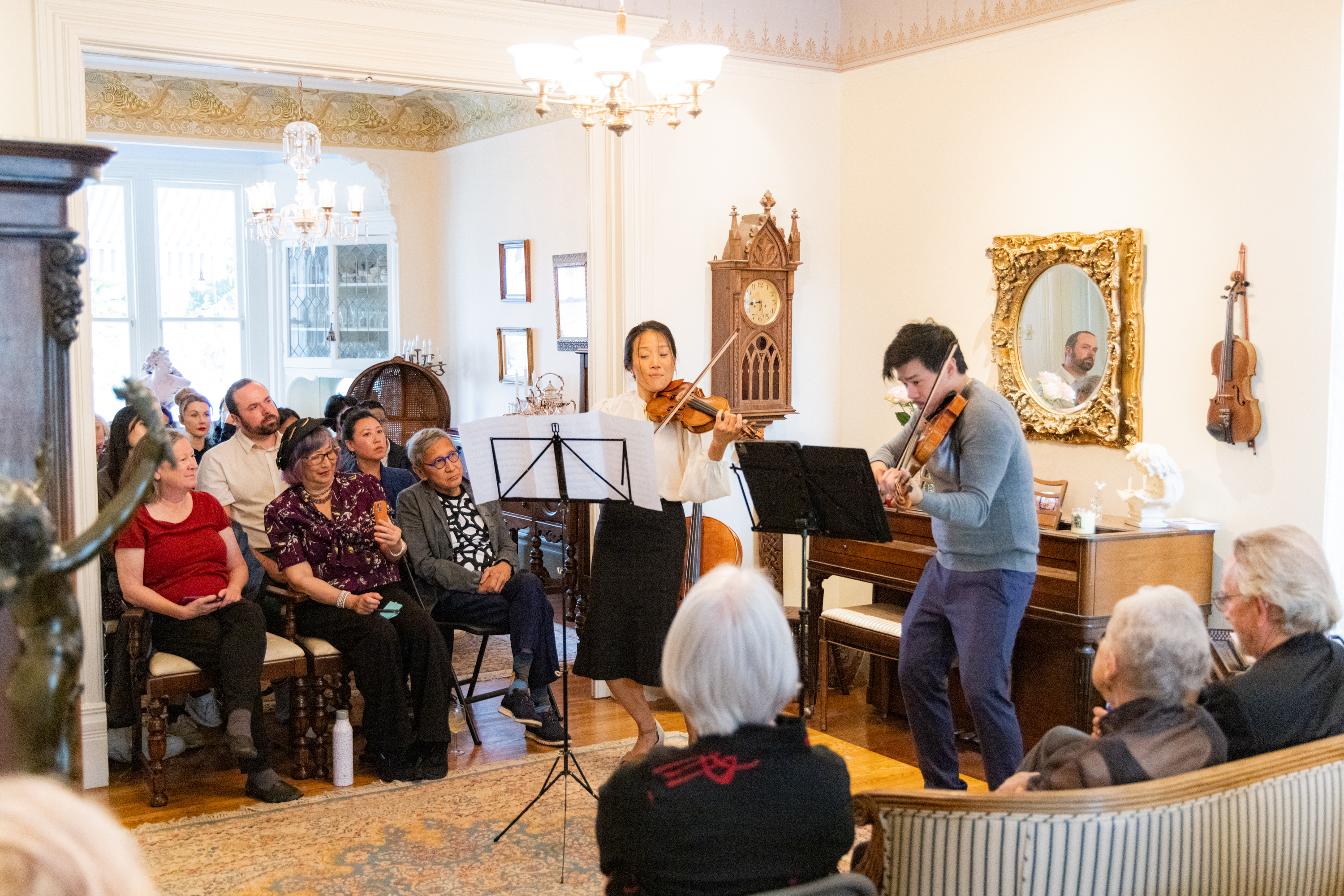 Two violinists perform in a cozy, well-decorated room as a small audience attentively listens, seated on chairs and a sofa.