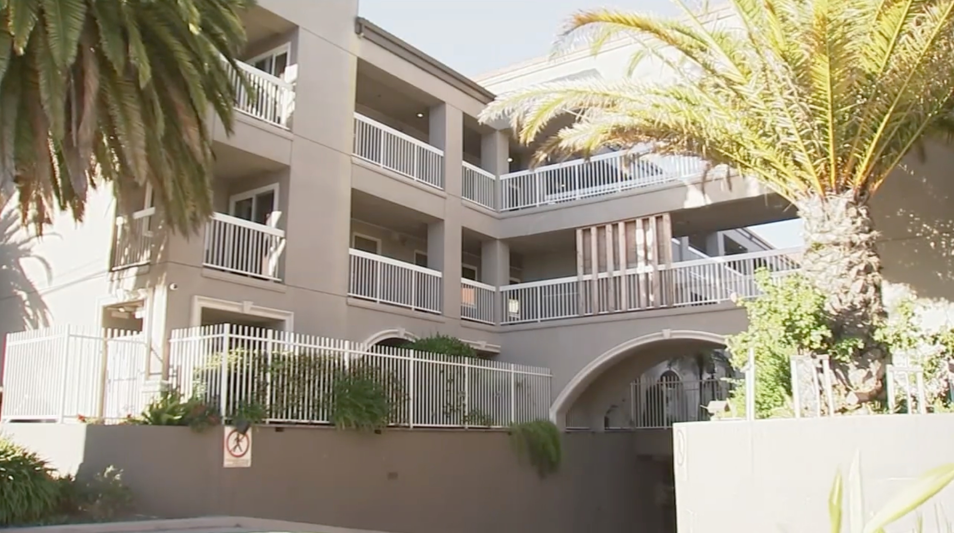 The image shows a multi-story residential building with balconies and white railings, surrounded by palm trees and greenery, with a &quot;No Pedestrians&quot; sign visible.