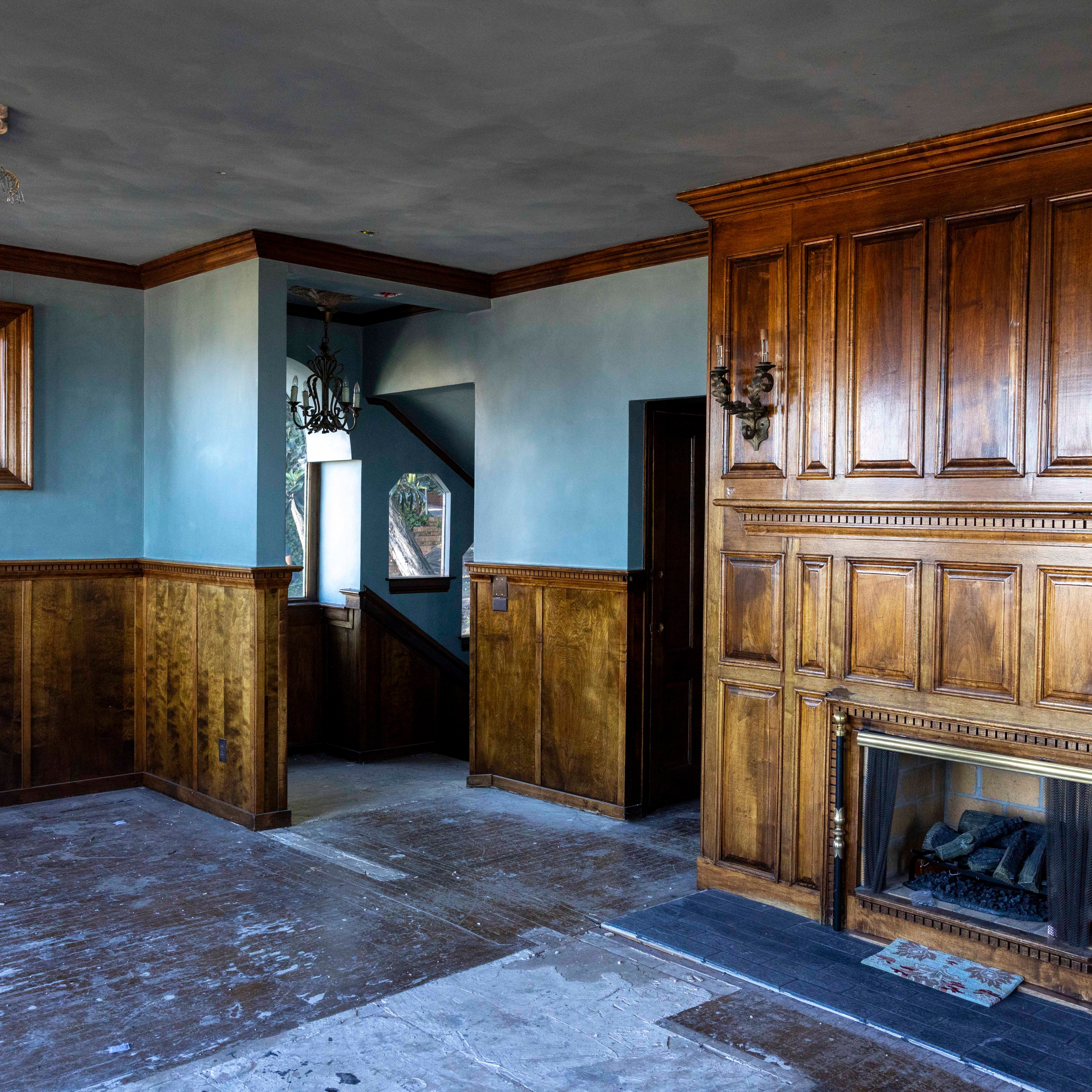 The image shows a room with blue walls and wooden wainscoting, a fireplace with wooden paneling, and a staircase with a chandelier visible through an archway. The floor appears unfinished.