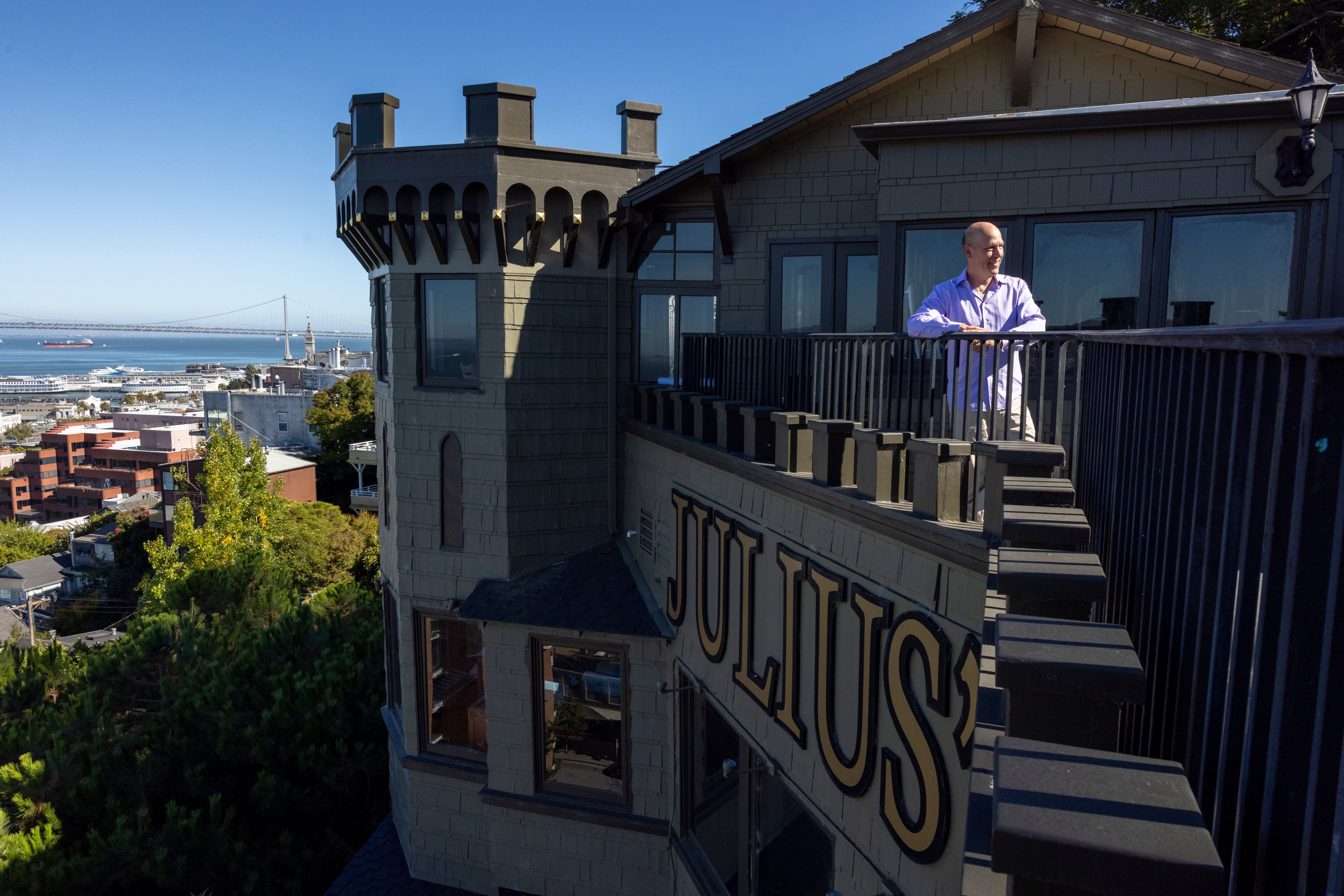 A man stands on a balcony with "Julius'" inscribed below him, overlooking a cityscape with a distant bridge and water visible under a clear blue sky.