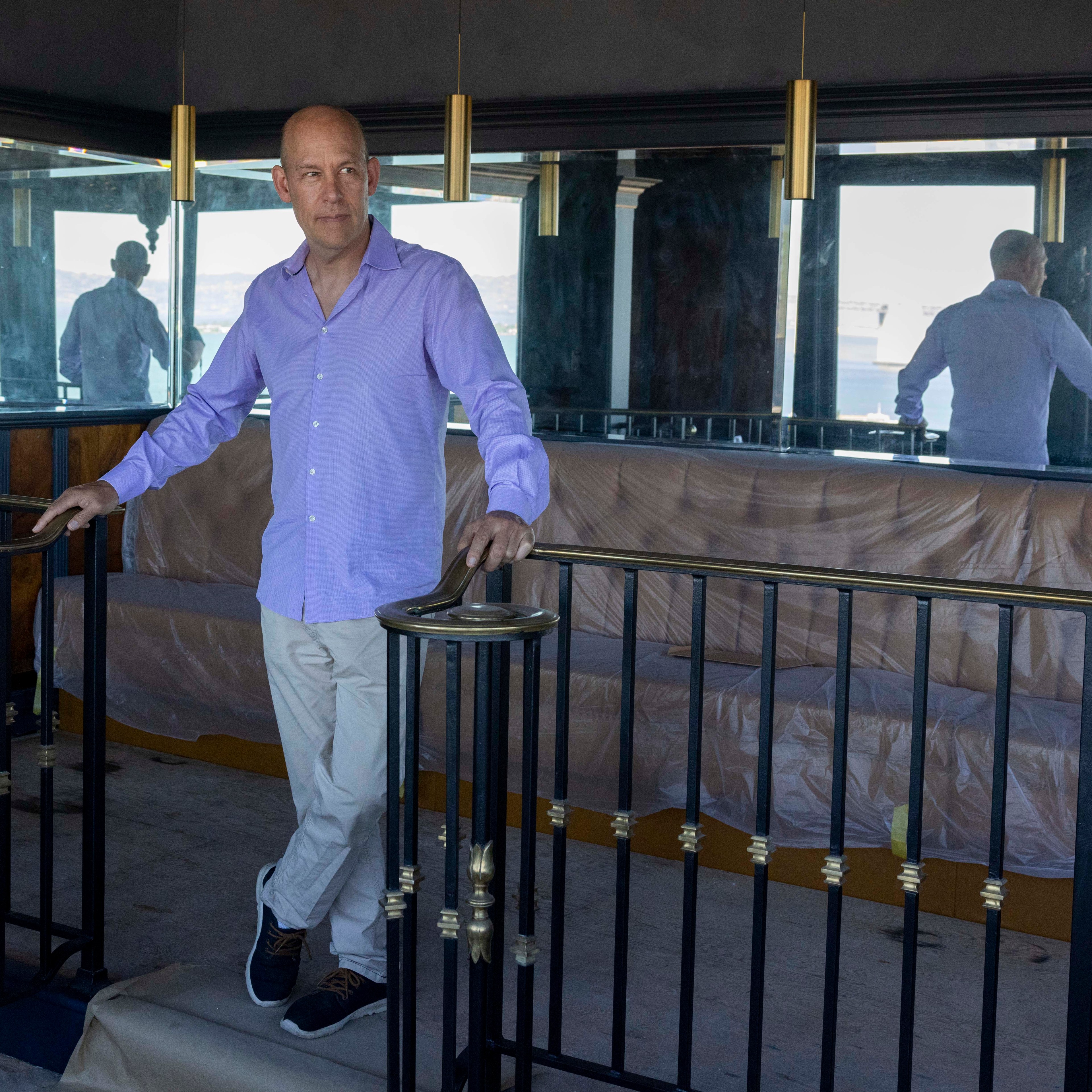 A man in a light purple shirt and beige pants stands by a black railing in front of a mirrored wall, with a plastic-covered couch and pendant lights in the background.