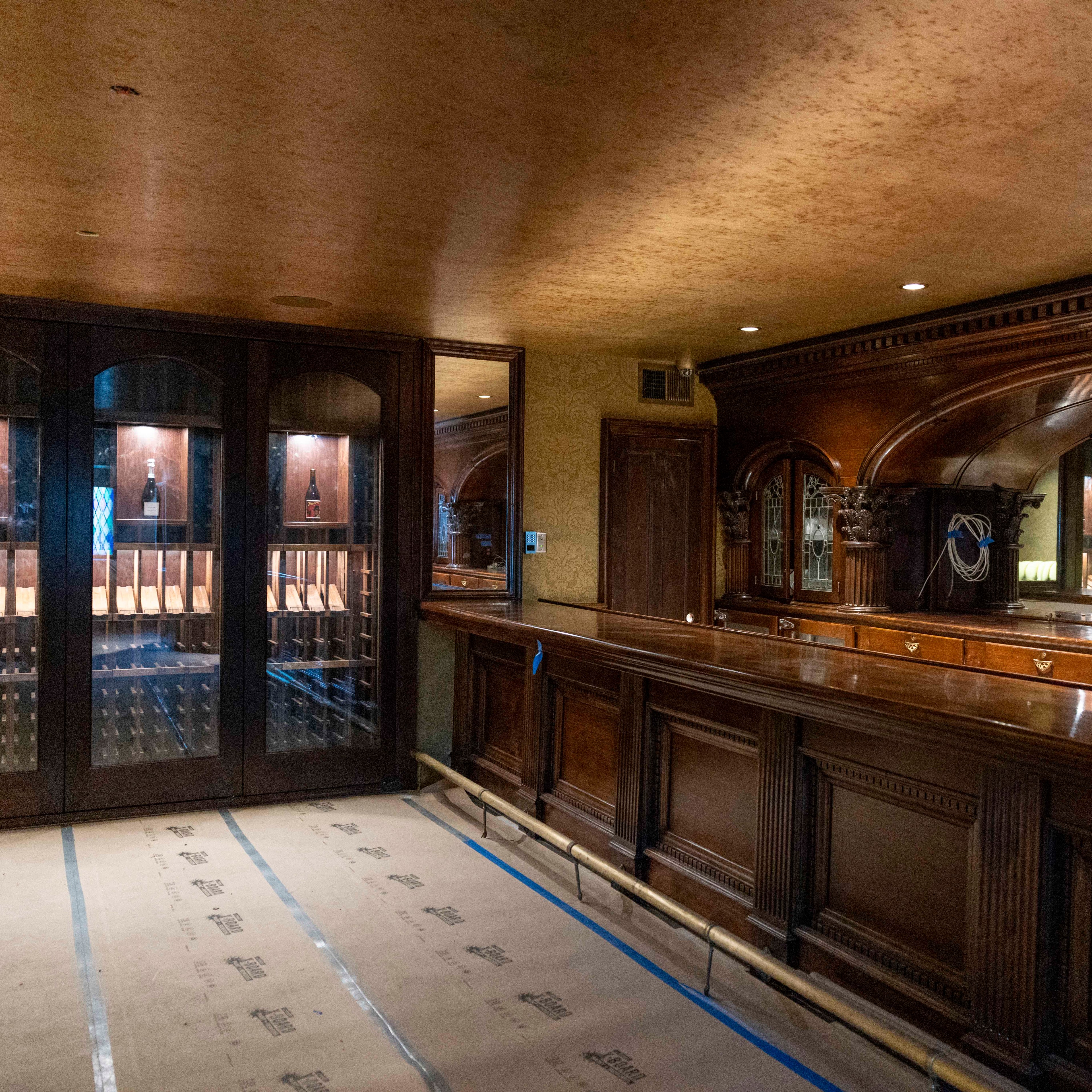 The image shows a room with rich, dark wood paneling, a long wooden bar, and a wine display cabinet with glass doors. The floor is covered with protective paper.