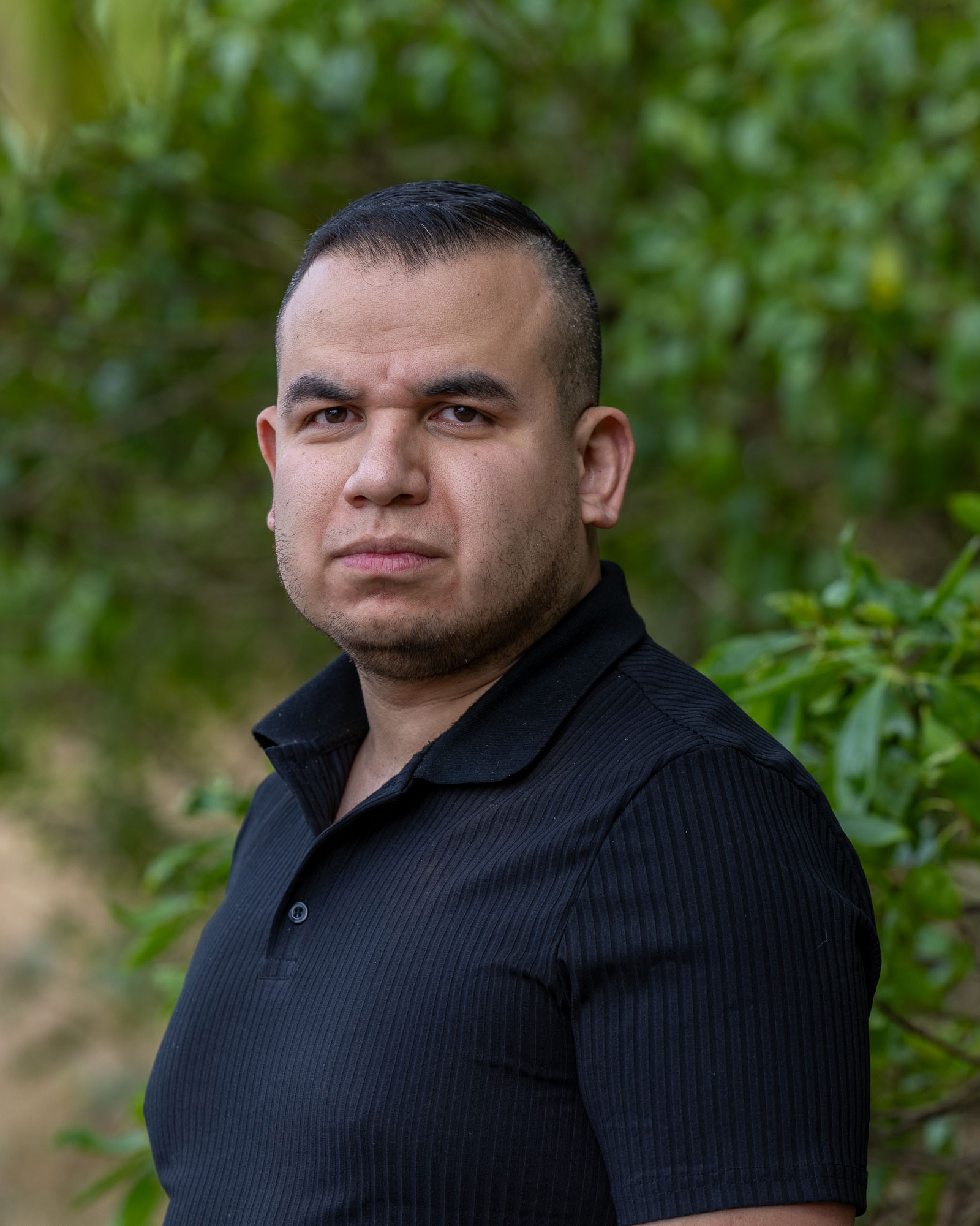 A man with short dark hair and a trimmed beard is wearing a black shirt and standing against a blurred green leafy background, looking directly at the camera.