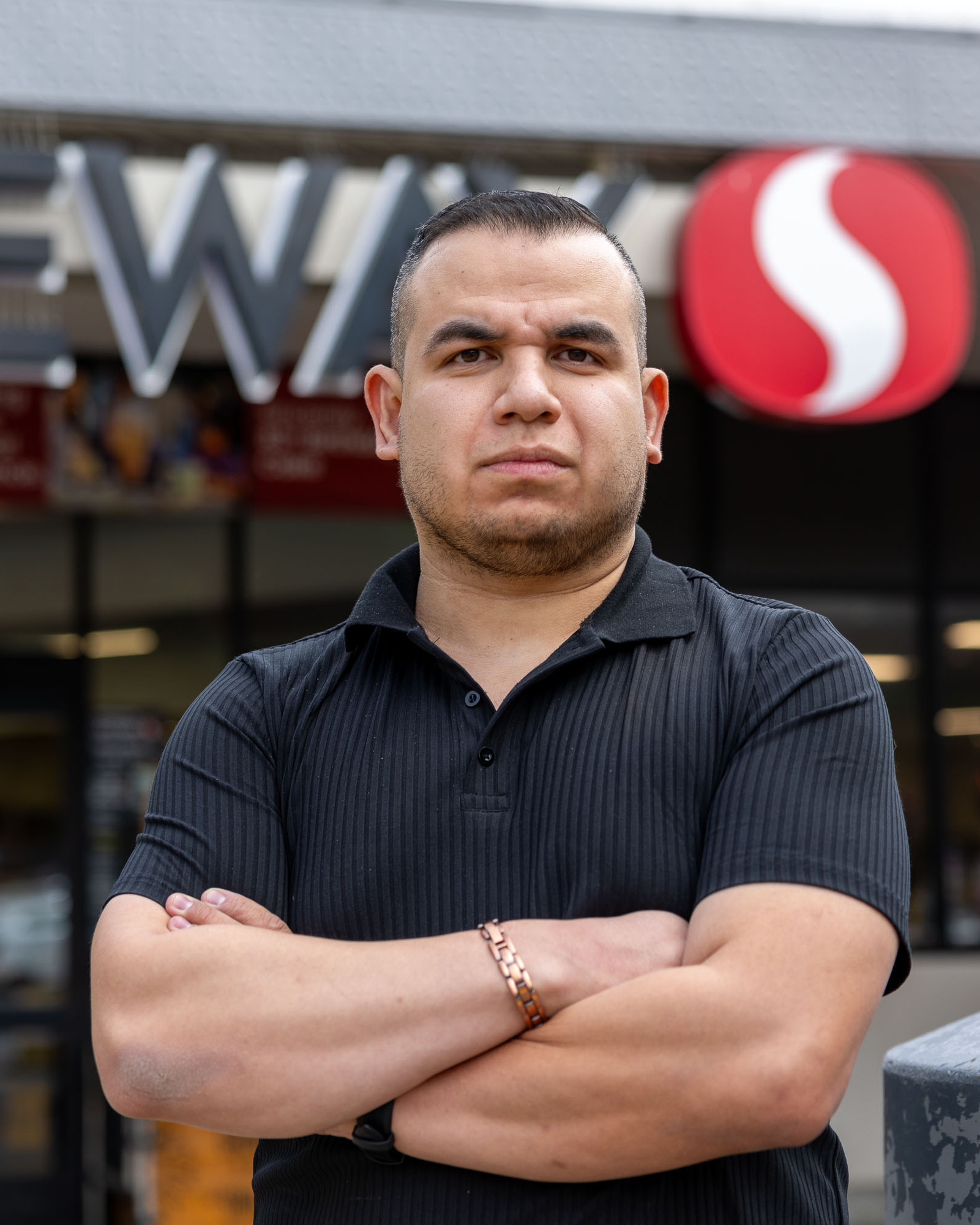 A man wearing a black shirt stands with his arms crossed in front of a store with a red and white logo above the entrance. He looks serious.