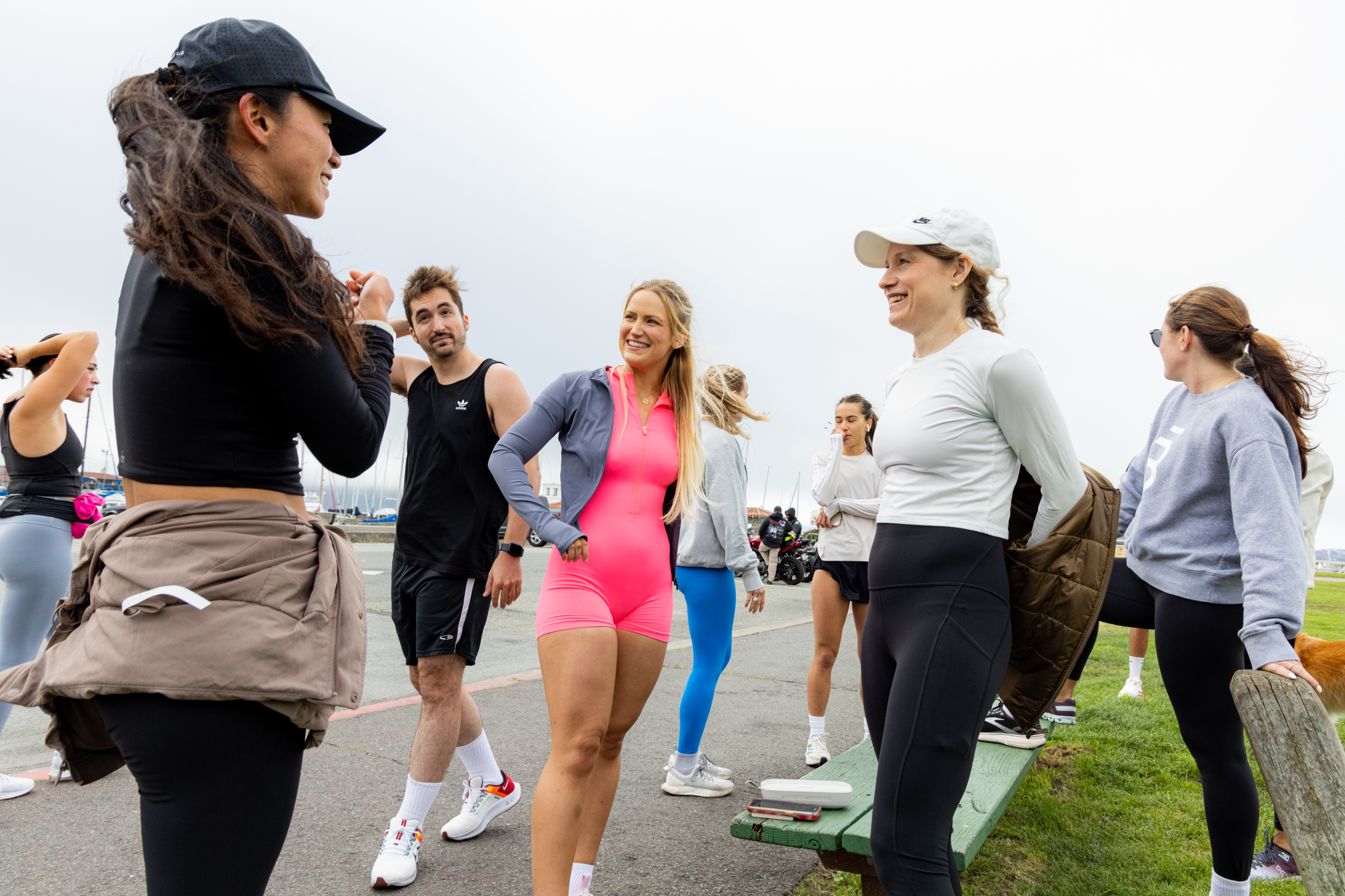 A group of people in athletic wear are gathered outdoors, some chatting and others stretching, with a background of a cloudy sky and water. One woman wears a bright pink outfit.