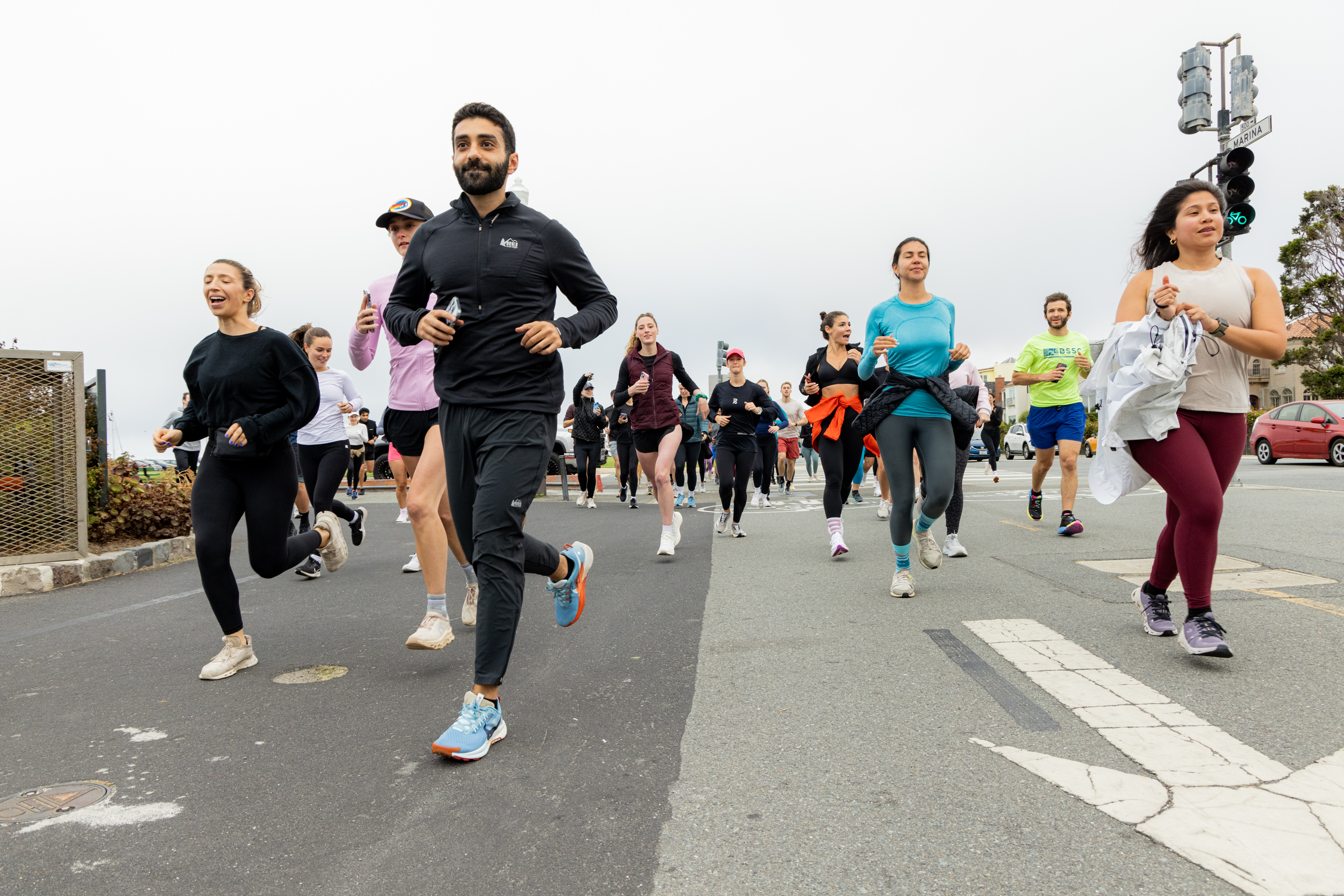 A group of people is jogging on a road, some smiling and others focused, all wearing athletic clothing. The background shows an overcast sky and traffic lights.