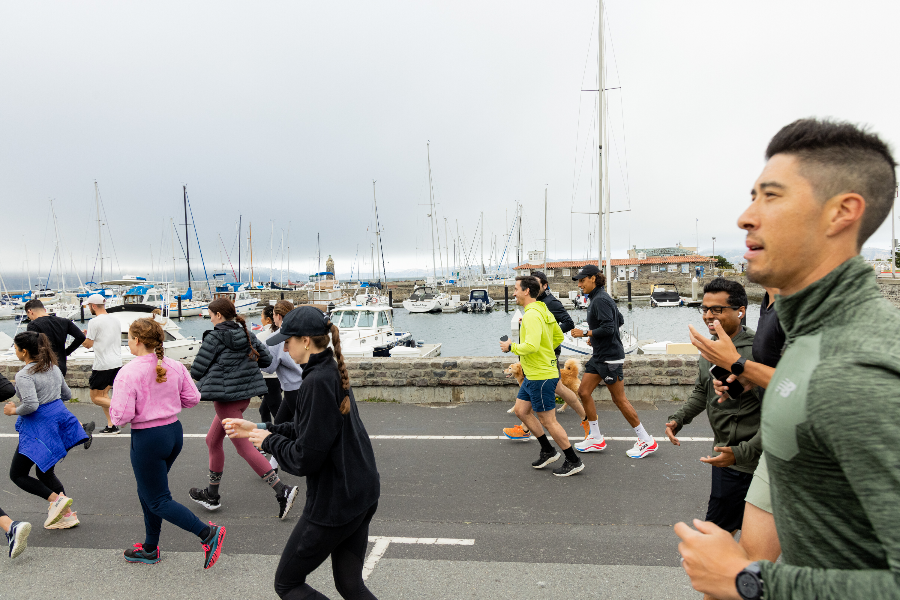 A group of people jogs near a marina filled with boats. They wear athletic clothing, and the overcast sky casts a gray hue over the scene.