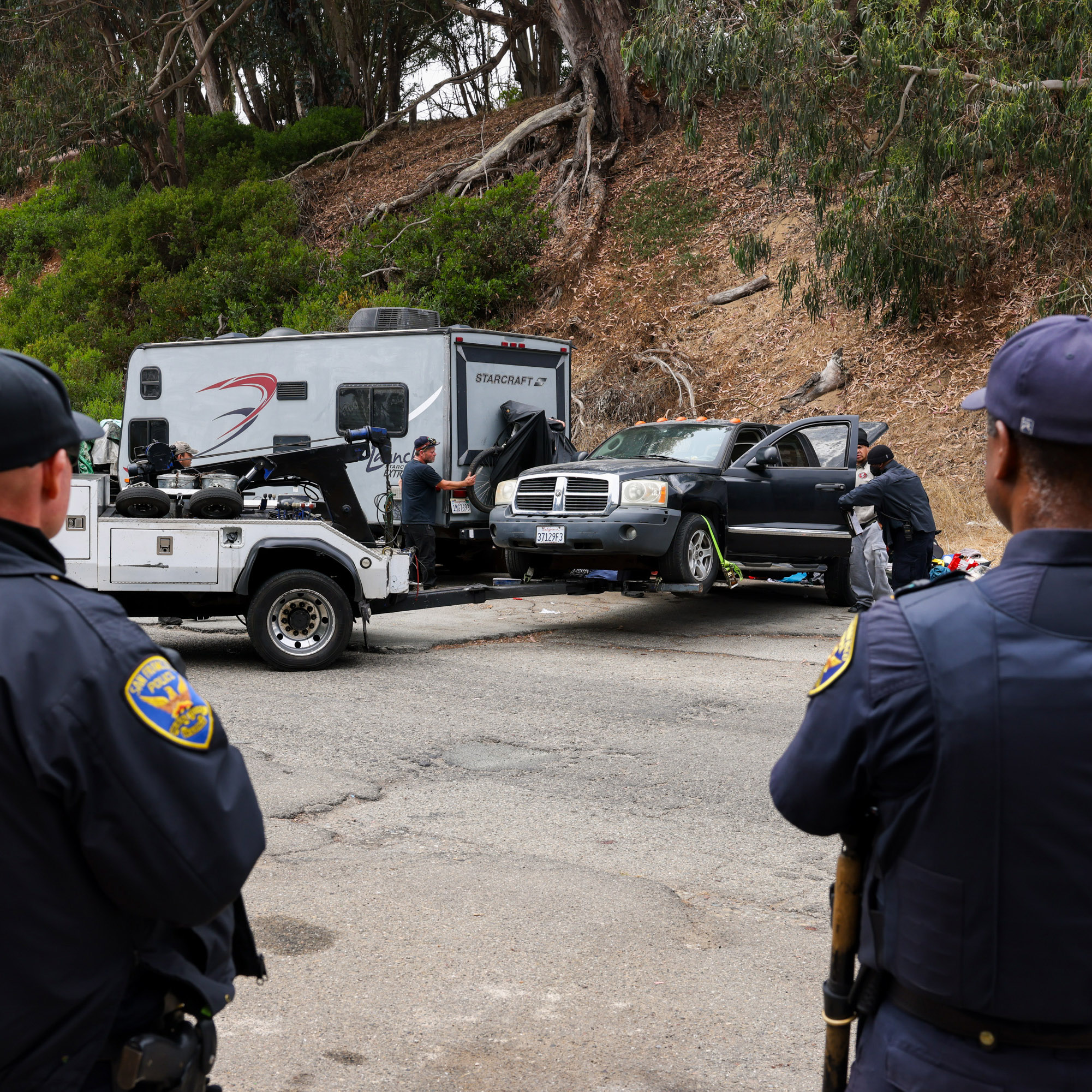 Police officers look on as two individuals, next to a black truck, work near an RV and a tow truck on a tree-lined roadside.