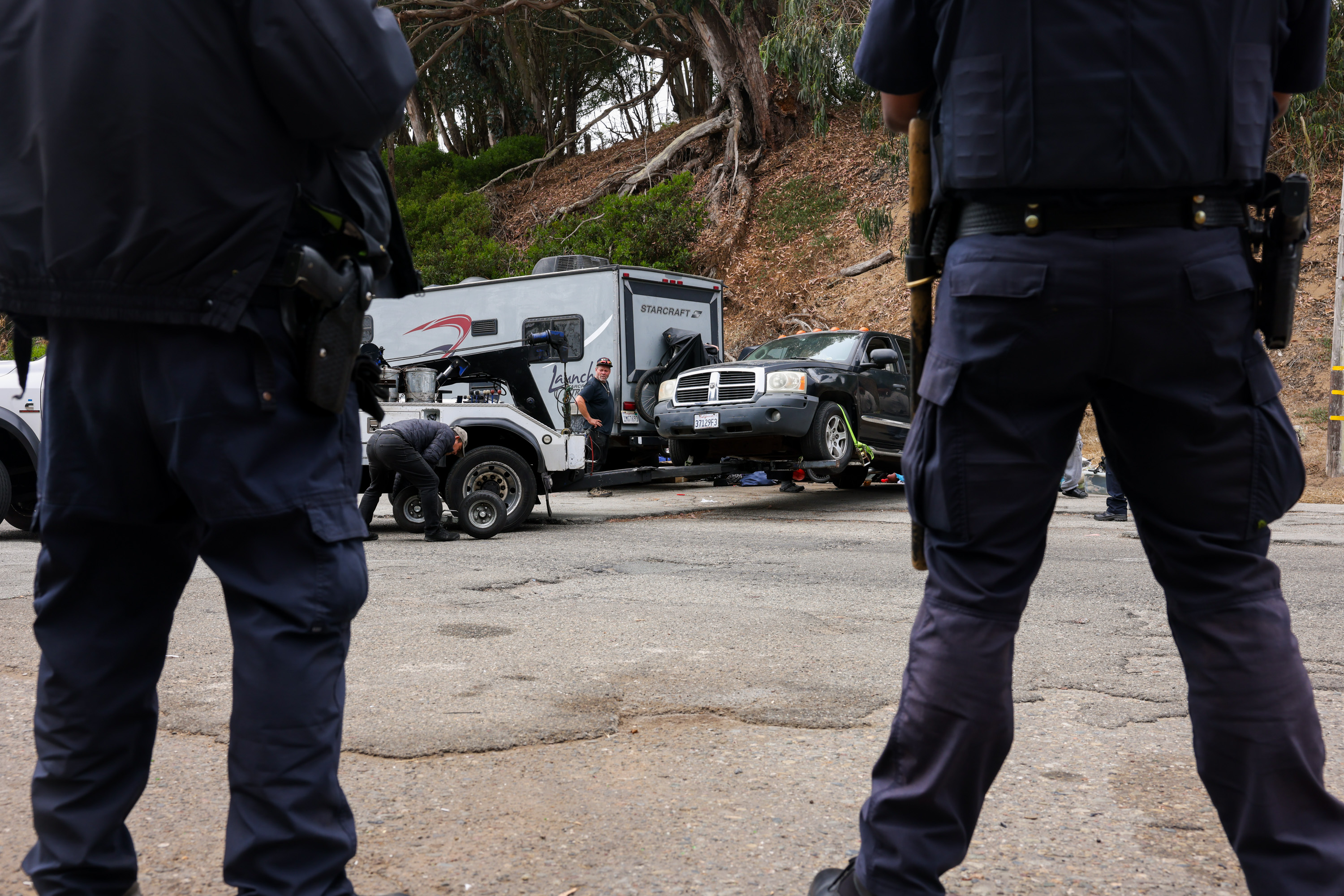 Two officers in uniform observe a scene involving a black SUV being worked on by mechanics, near a tow truck and an RV, with trees and hills in the background.