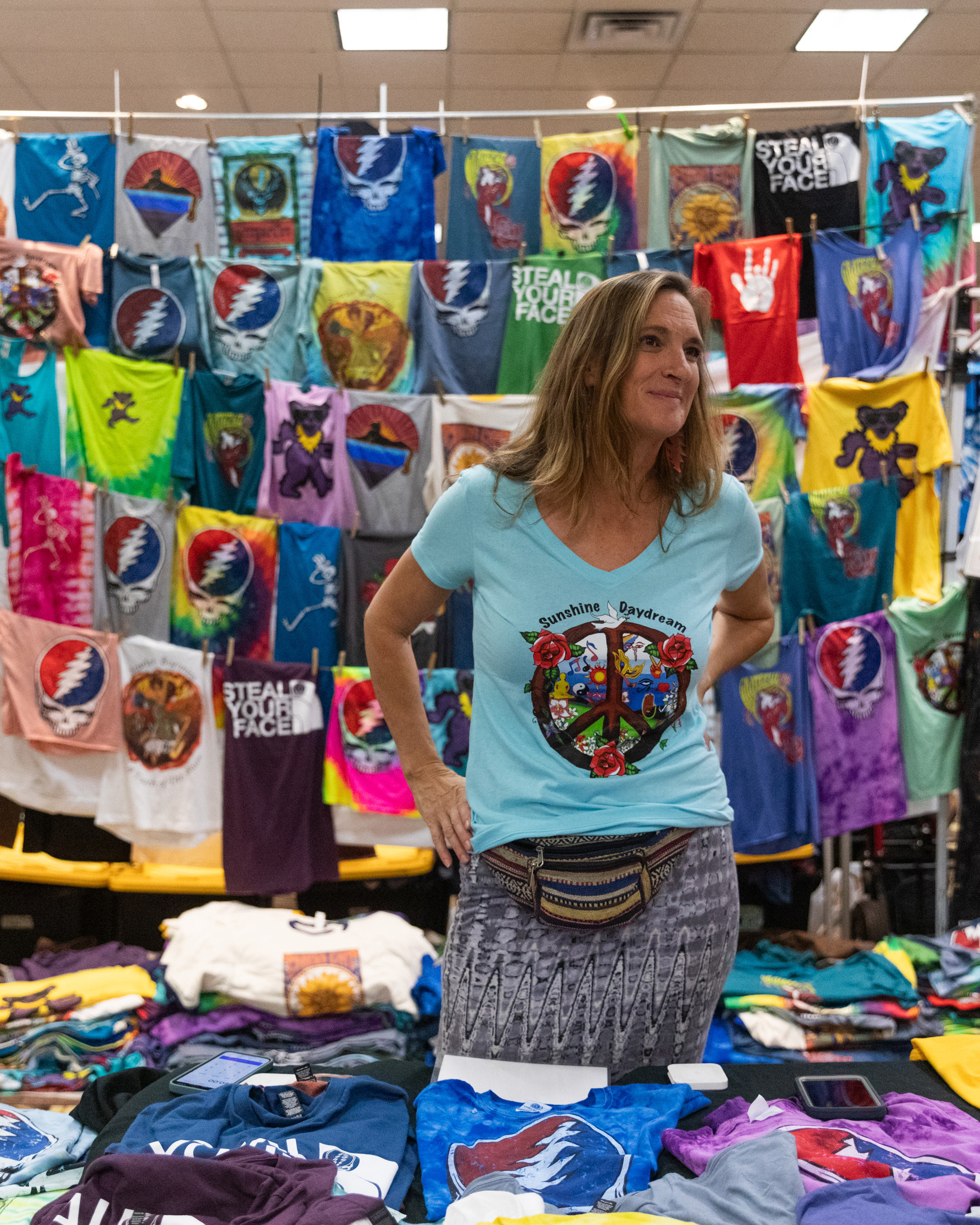 A woman stands at a booth smiling, surrounded by colorful, Grateful Dead-themed tie-dye t-shirts hanging on display and neatly folded on tables.