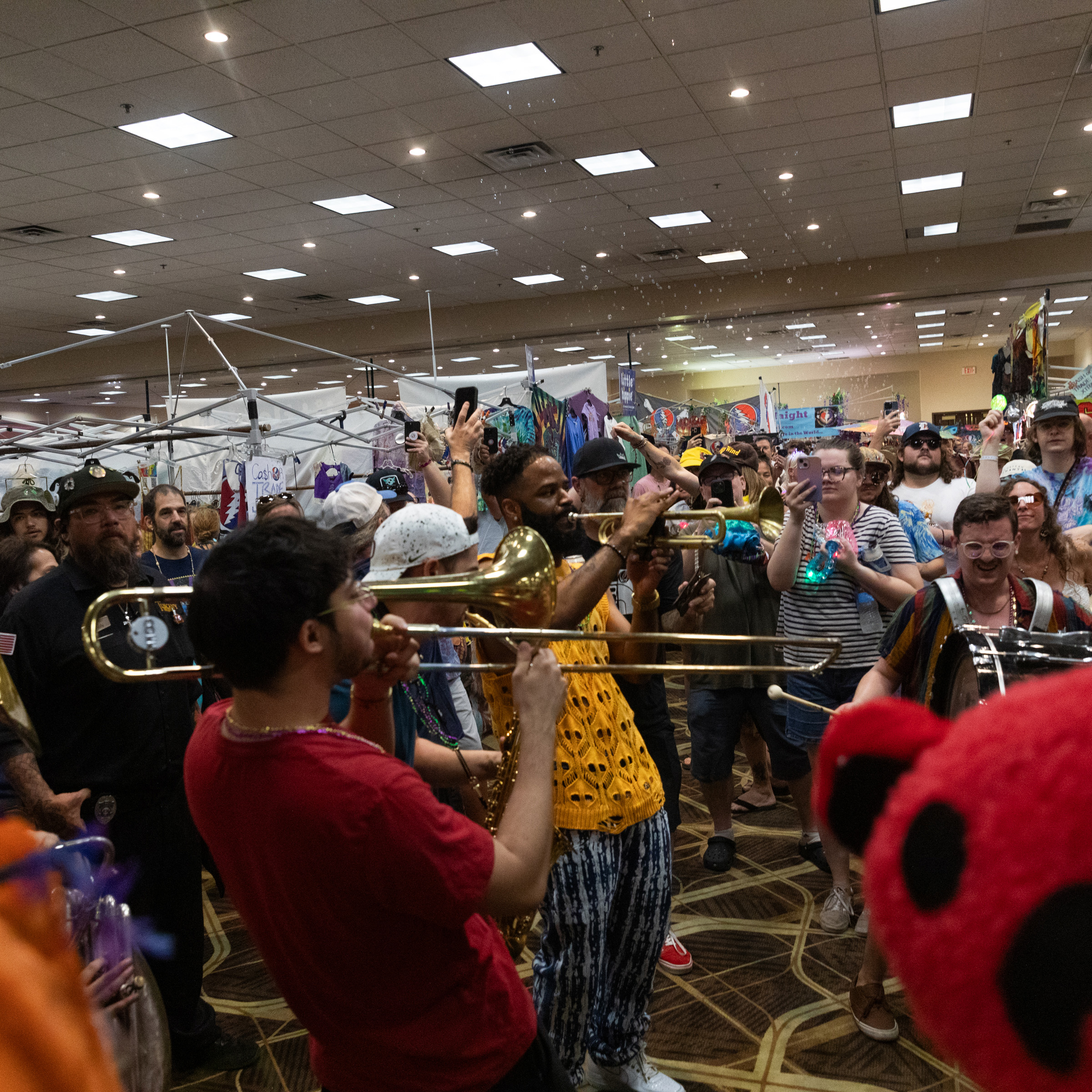 A lively crowd watches a brass band play music indoors, with people recording on their phones, surrounded by various merchandise booths and festive decor.