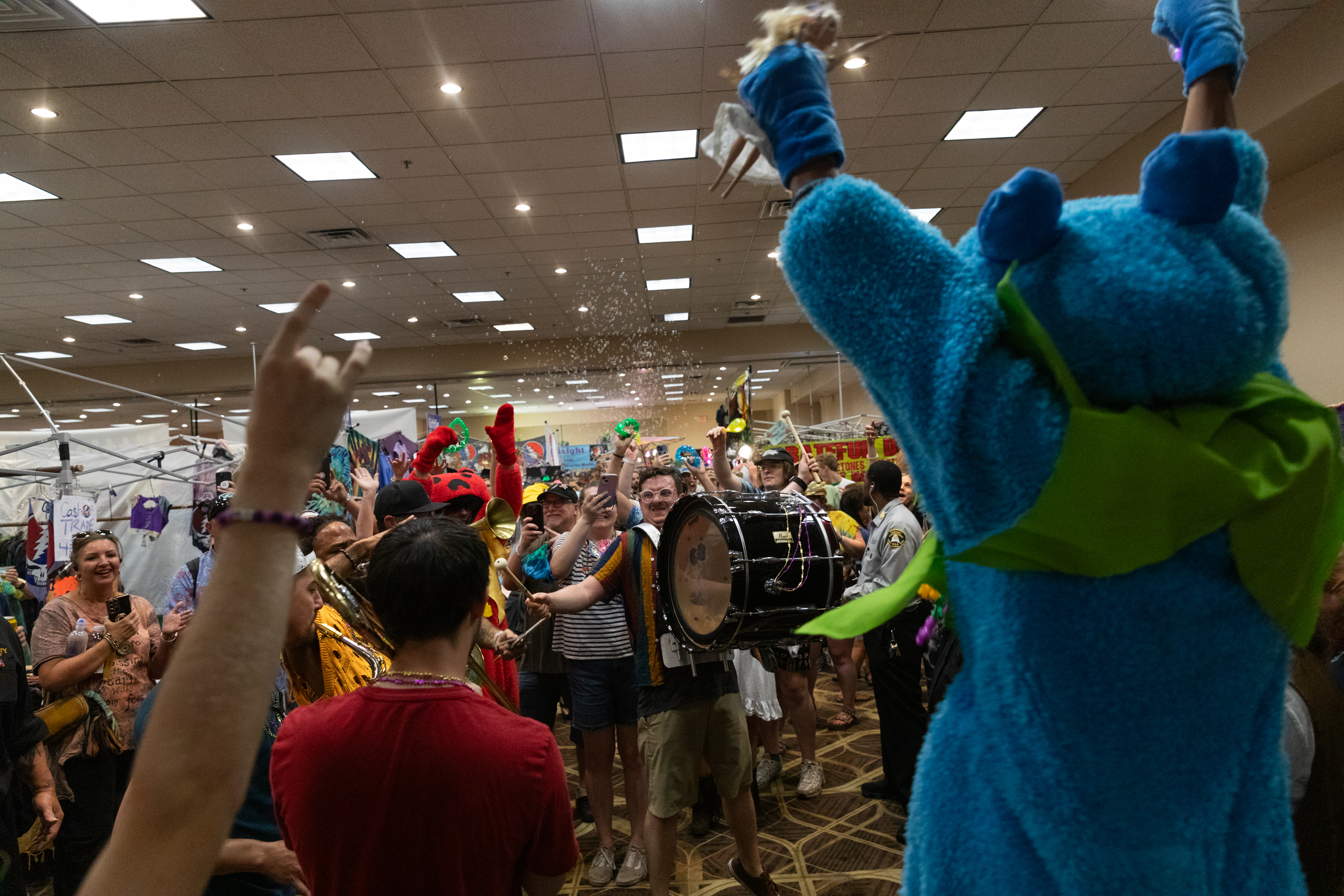 A person in a blue animal costume interacts with a lively crowd at an indoor event, featuring confetti, musical instruments, and colorful decorations.