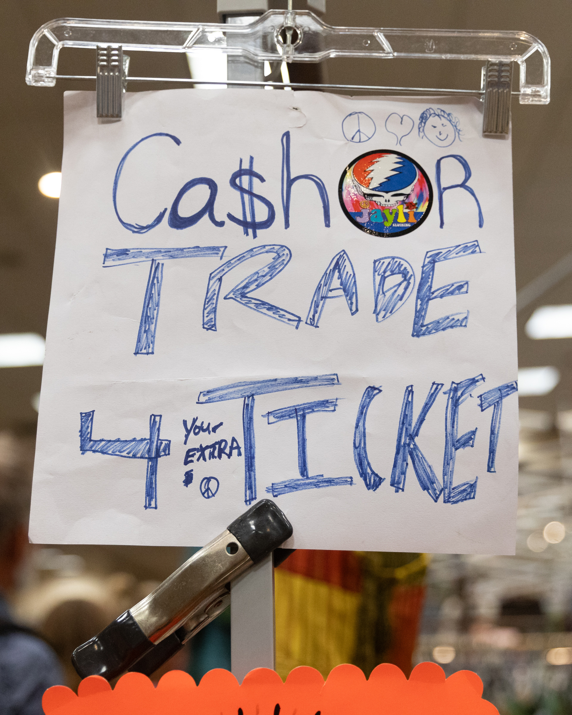 A hand-drawn sign on a hanger reads &quot;Cash or Trade 4 your extra Ticket&quot; with peace signs and a colorful sticker.