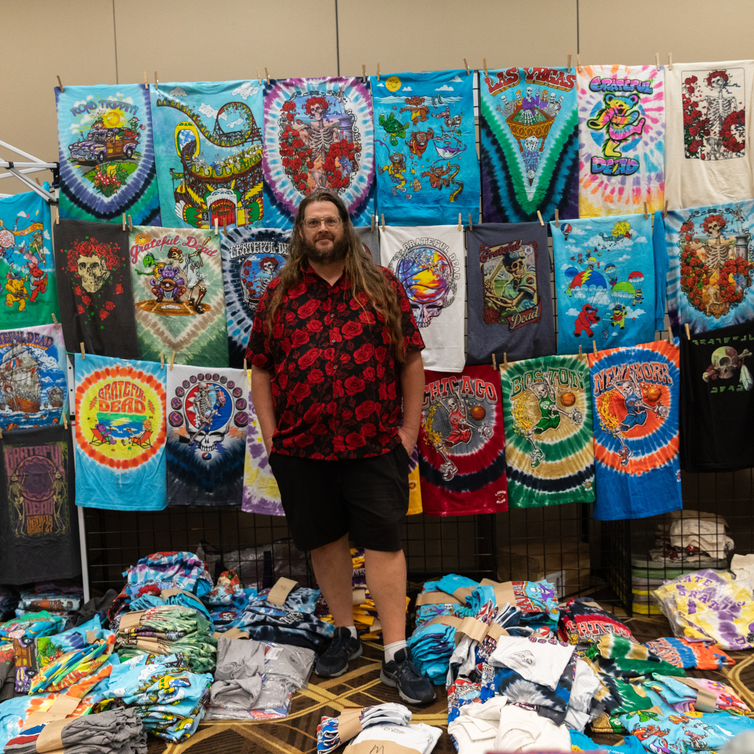 A person stands in front of a colorful display of tie-dye and graphic T-shirts, with more shirts piled on the floor. They wear a red rose-patterned shirt and black shorts.