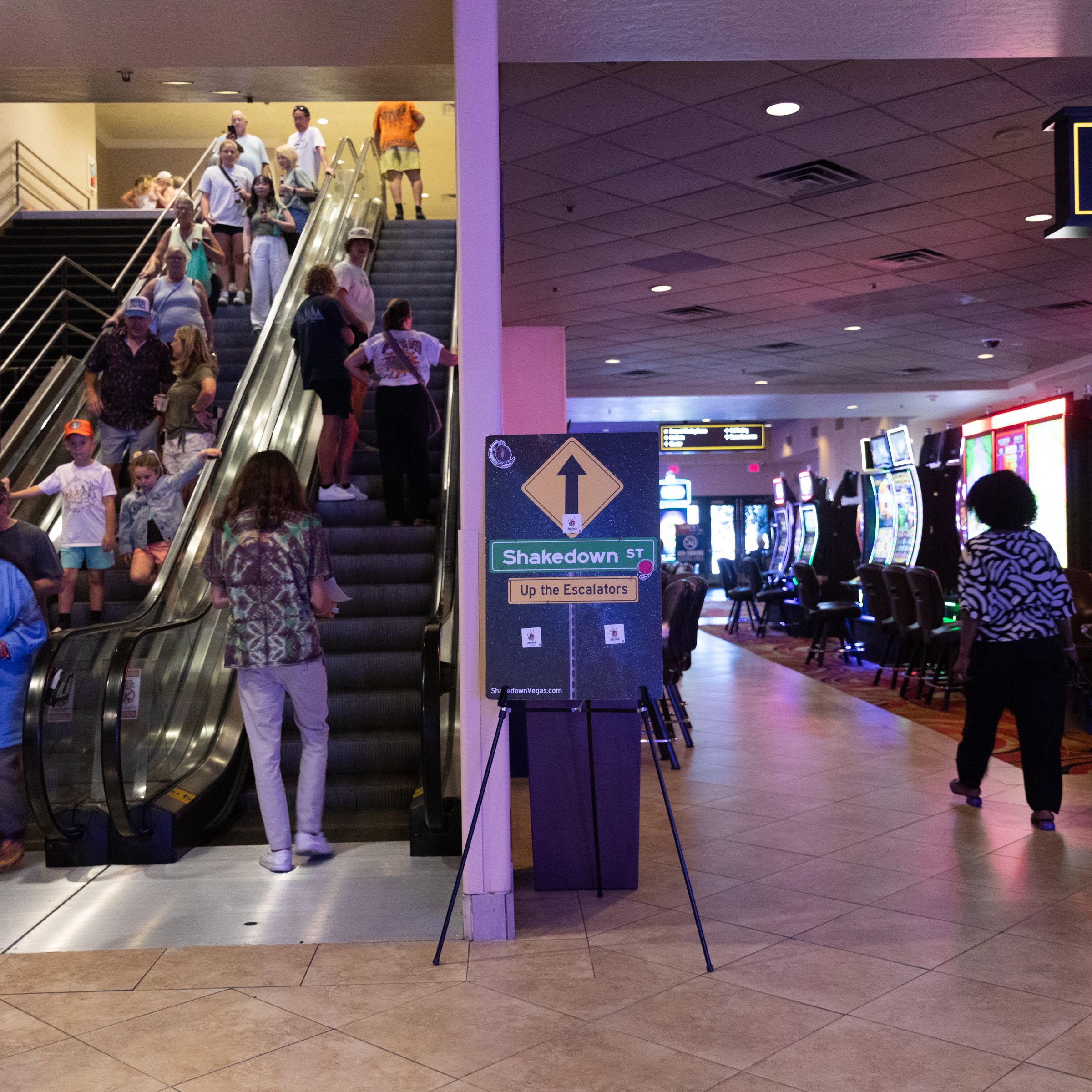 The image shows a group of people on escalators in a dimly lit indoor setting, with a sign saying &quot;Shakedown Street Up the Escalators&quot; and a casino area to the right.