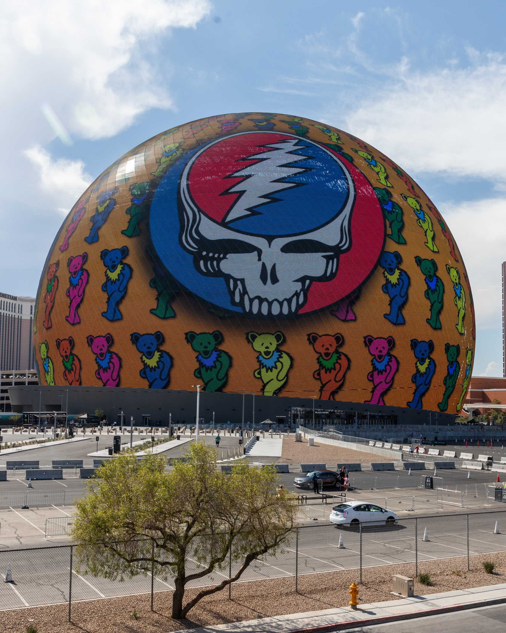 A large, round building displays a colorful graphic featuring a skull with a lightning bolt and dancing bears. The foreground includes a parking lot and a tree.