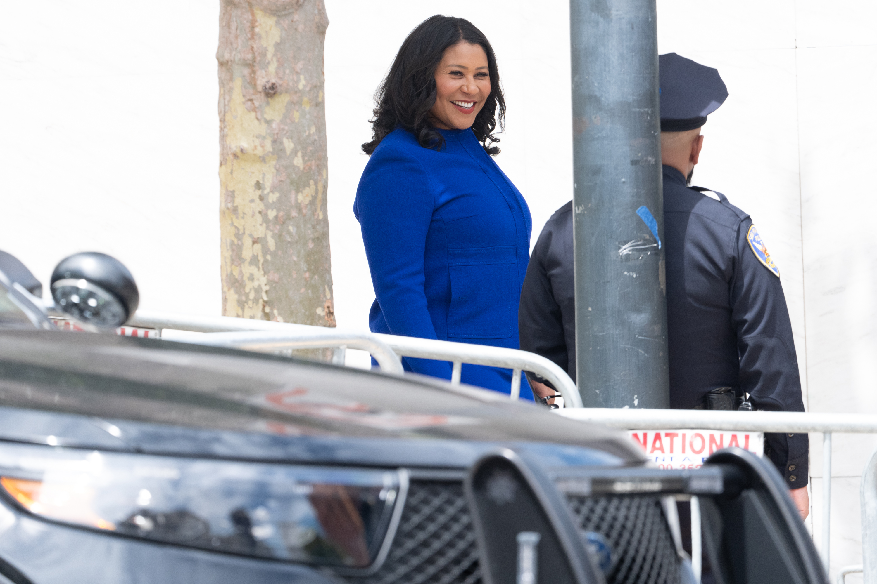 A woman in a bright blue outfit smiles while standing near a police officer in uniform. They are outside, next to a tree and a dark vehicle.