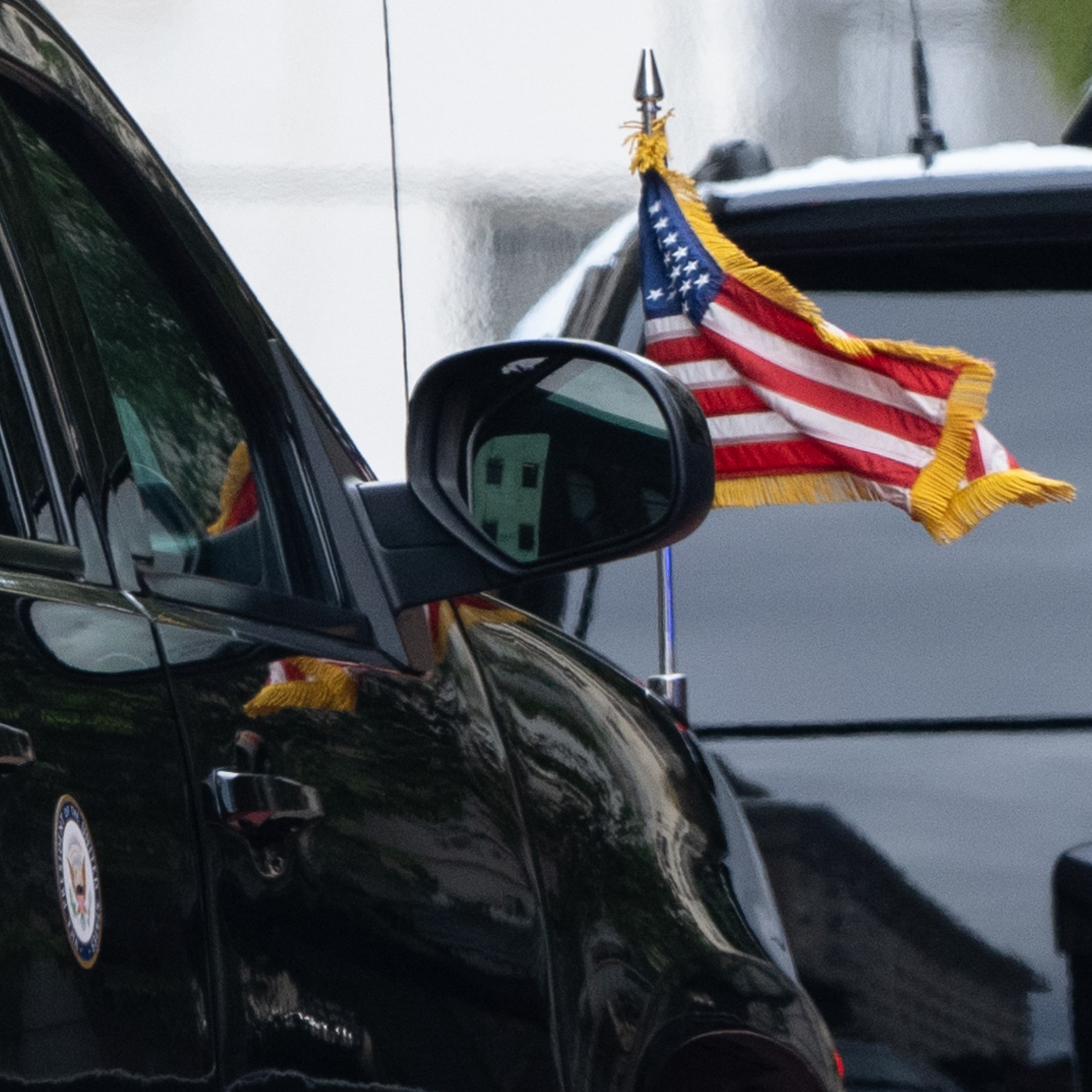 A black car with a side mirror displays a small American flag attached to it. Another vehicle's rear, with a visible brake light, is seen ahead.