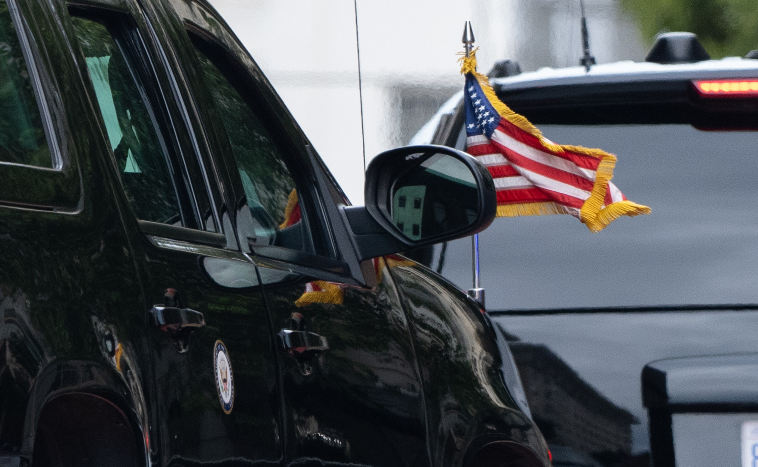 A black car with a side mirror displays a small American flag attached to it. Another vehicle's rear, with a visible brake light, is seen ahead.