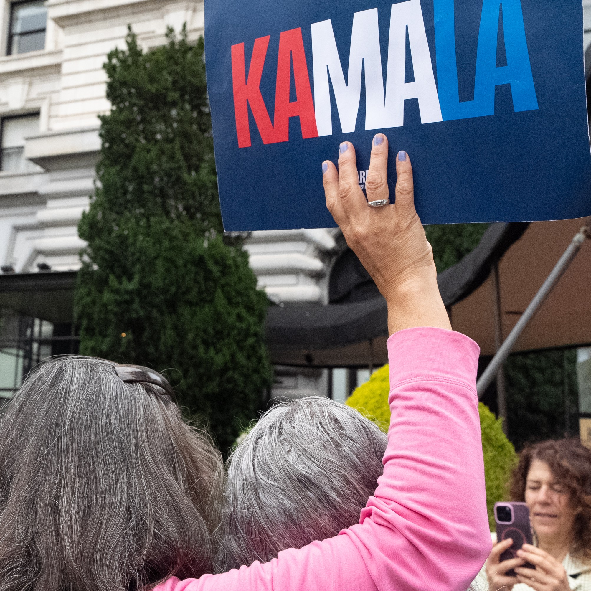 A crowd gathers in front of a large building, with a person holding a sign that reads &quot;KAMALA&quot; in red, white, and blue. Another person is taking a photo.