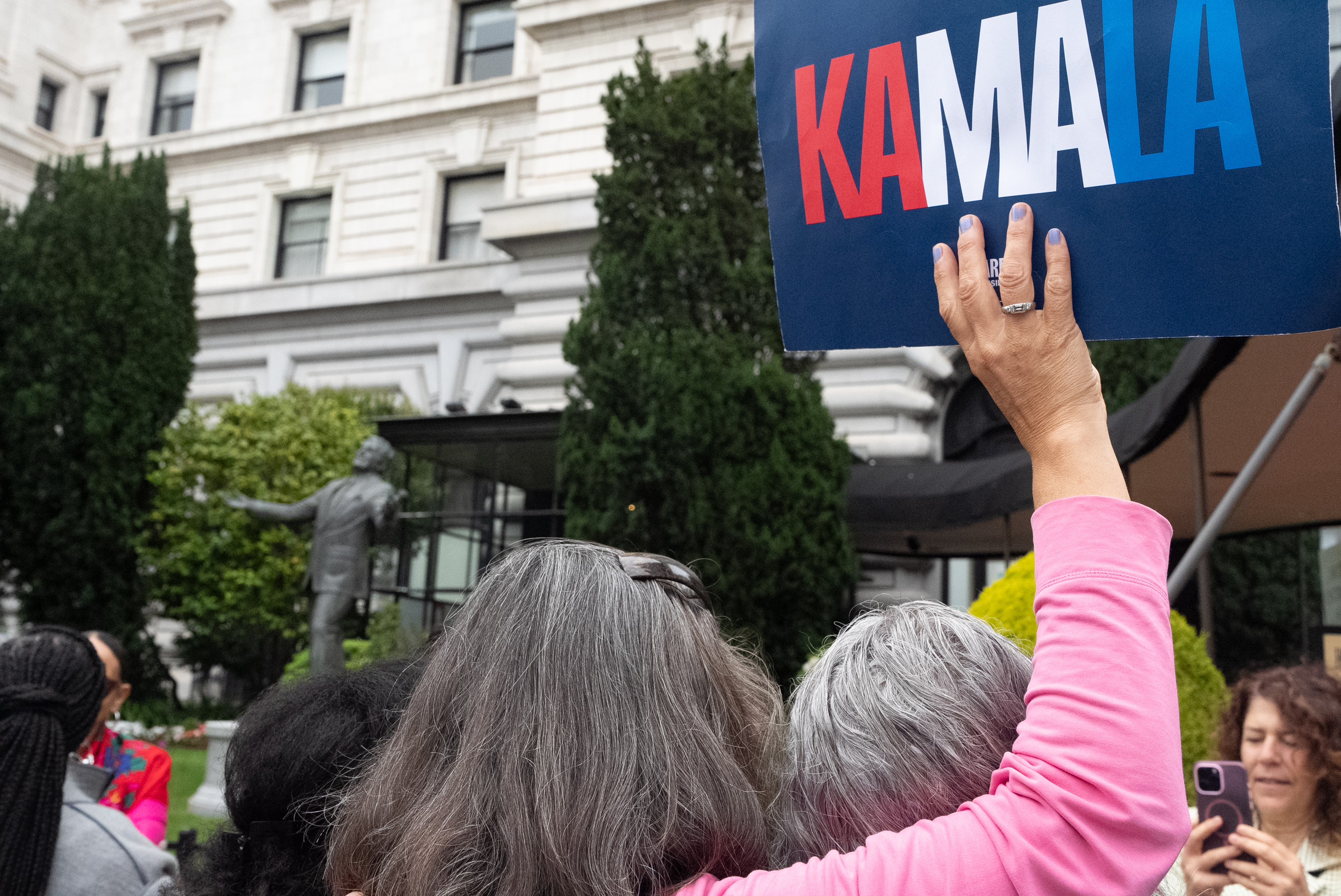 A crowd gathers in front of a large building, with a person holding a sign that reads &quot;KAMALA&quot; in red, white, and blue. Another person is taking a photo.