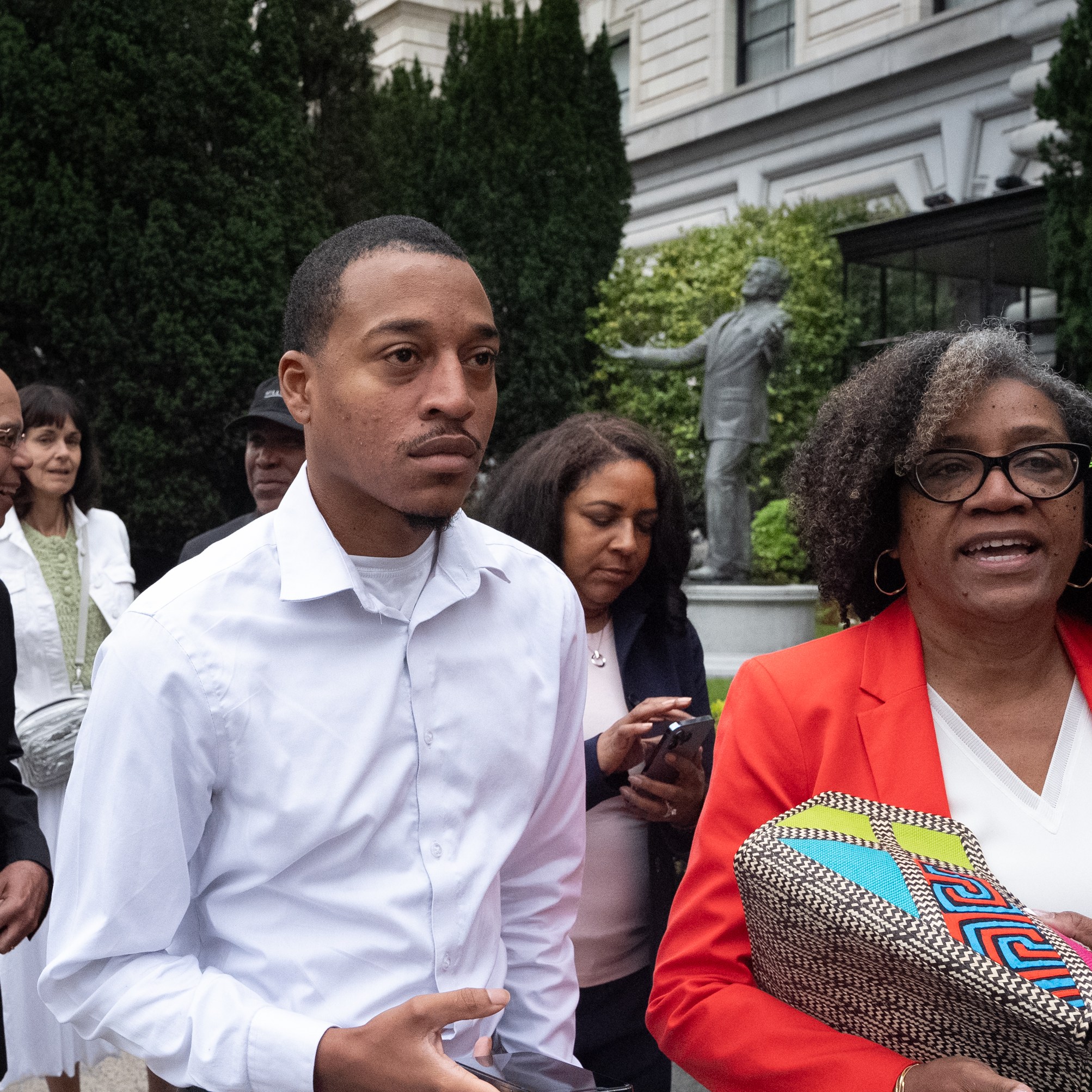 A group of people, dressed in business casual clothing, stand outdoors near a building with greenery in the background. They appear to be engaged in conversation.