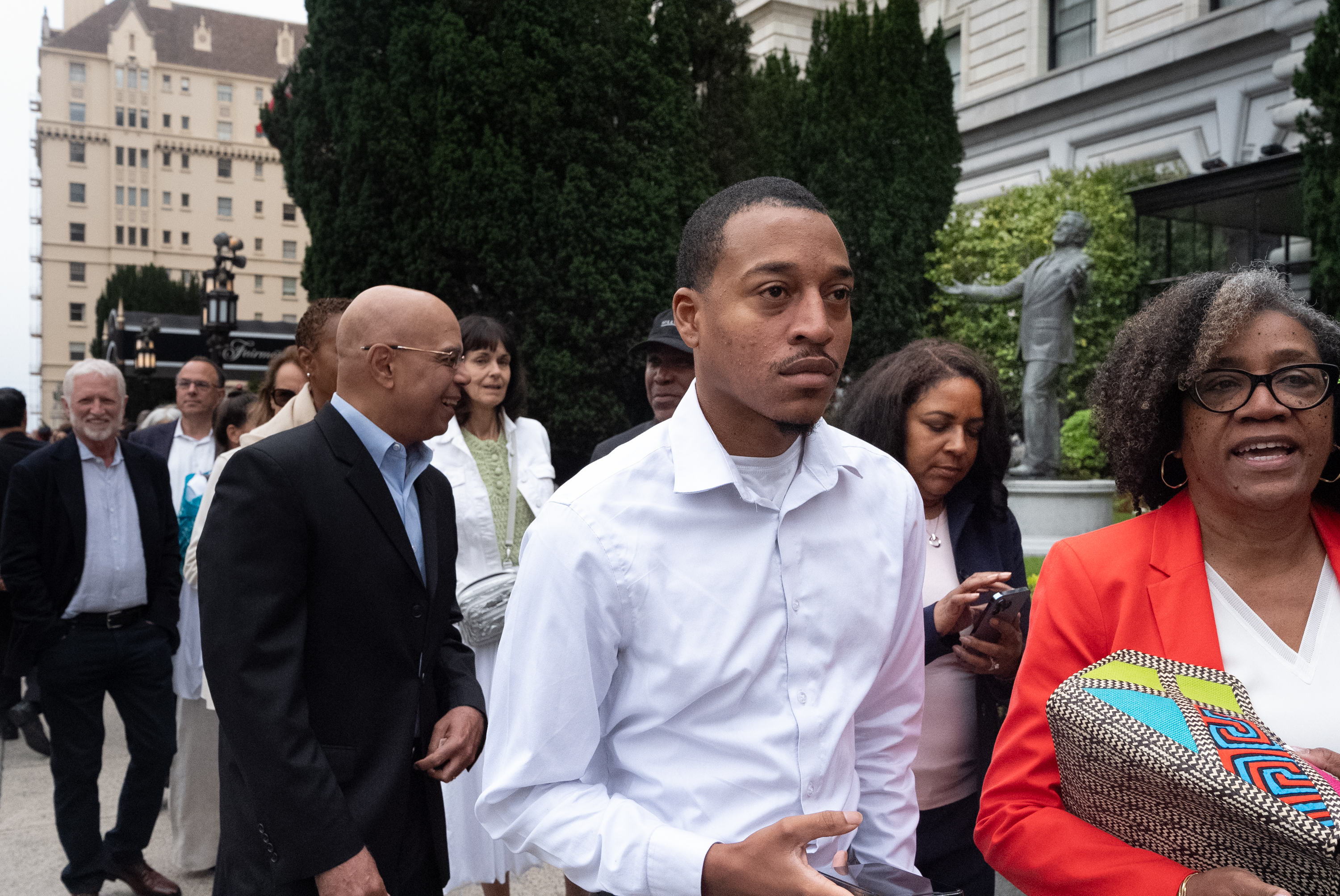 A group of people, dressed in business casual clothing, stand outdoors near a building with greenery in the background. They appear to be engaged in conversation.